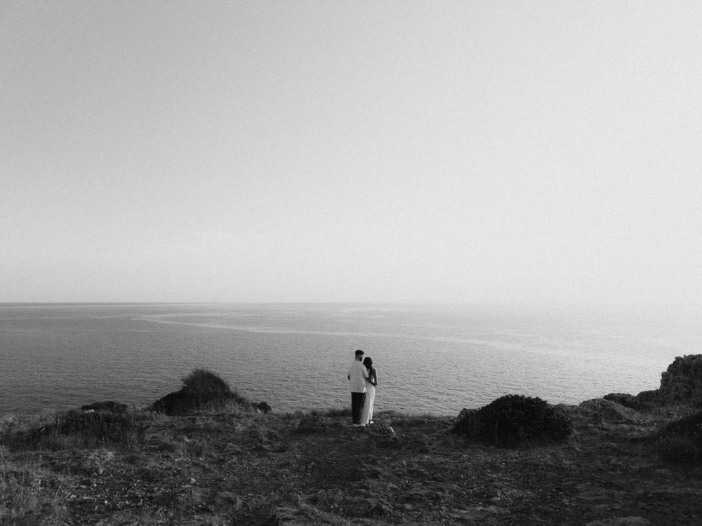 Black and white photo of a couple embracing on a rocky cliff overlooking a vast, calm ocean horizon.