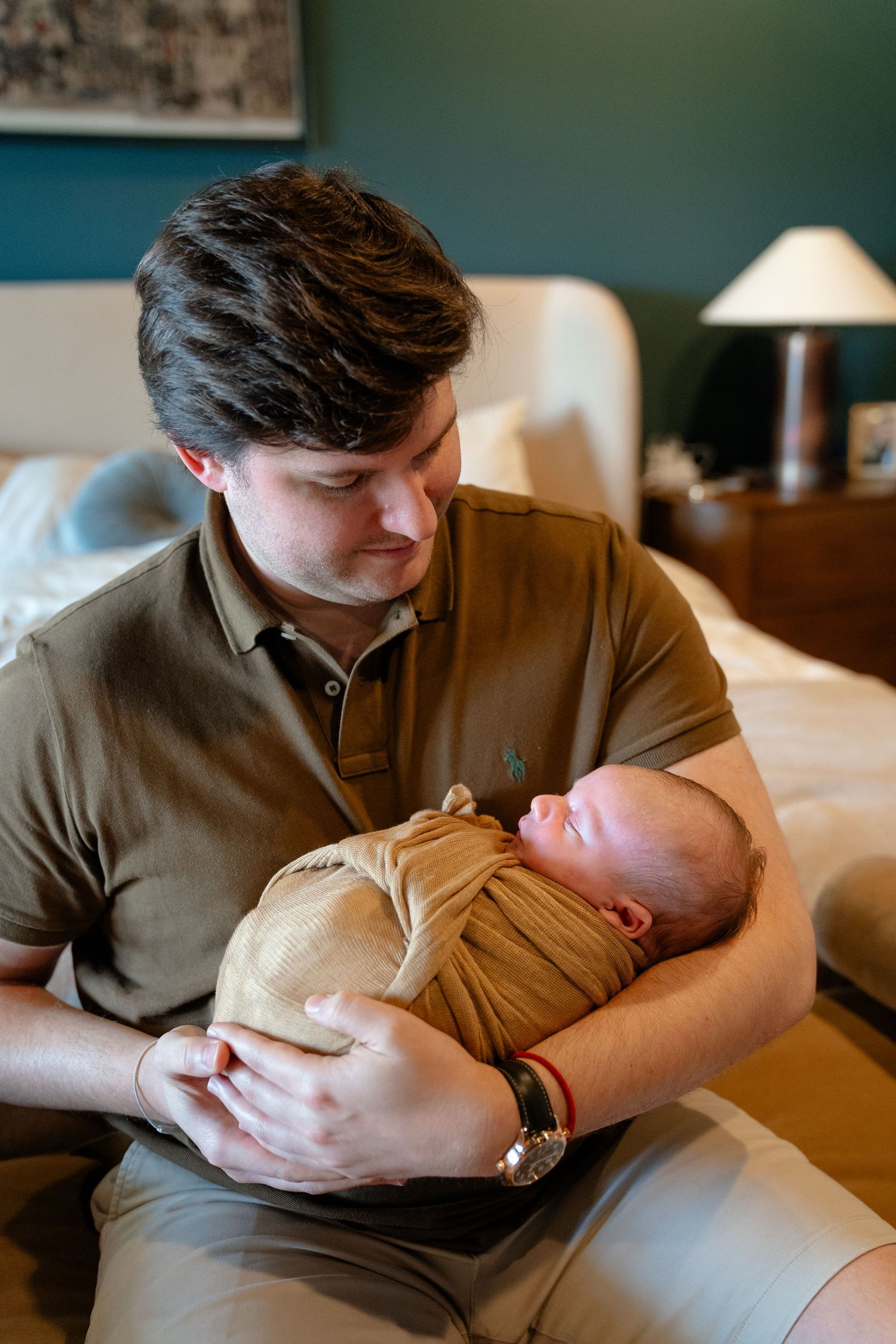 A man in a olive polo shirt tenderly holds a swaddled newborn baby while seated in a bedroom.