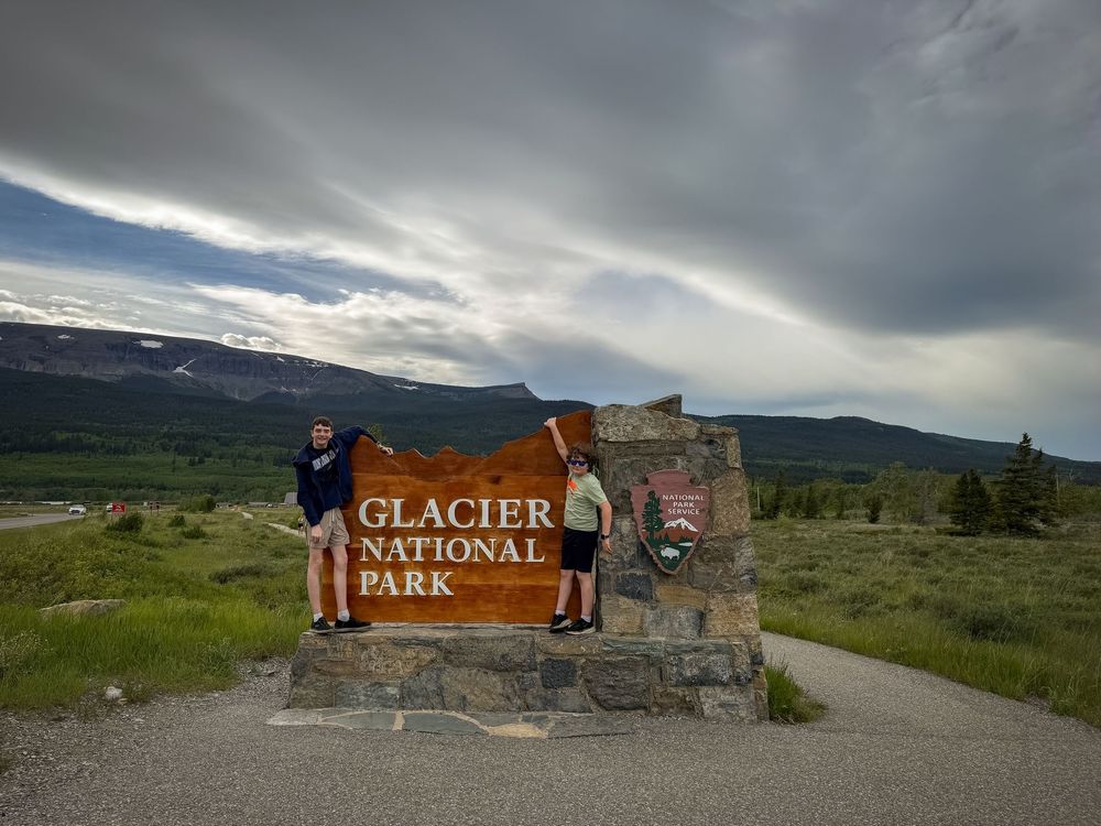 Two visitors pose beside the iconic Glacier National Park wooden entrance sign surrounded by mountains and dramatic cloudy skies.