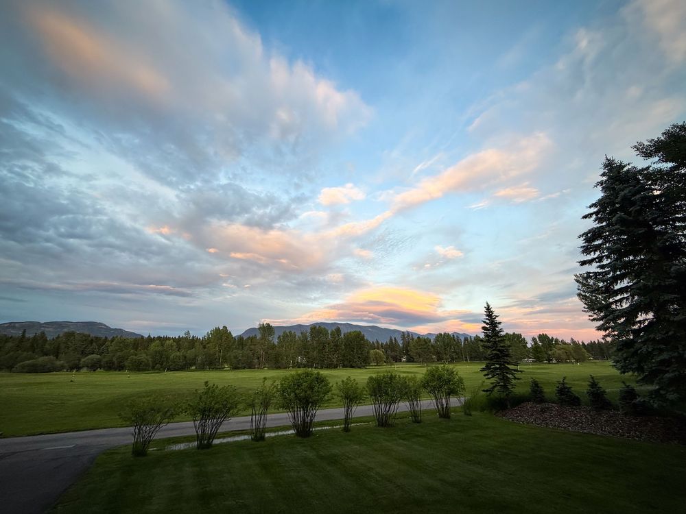 Scenic golf course at sunset with lush green fairways, tree-lined path, mountain backdrop, and colorful sky with wispy clouds.