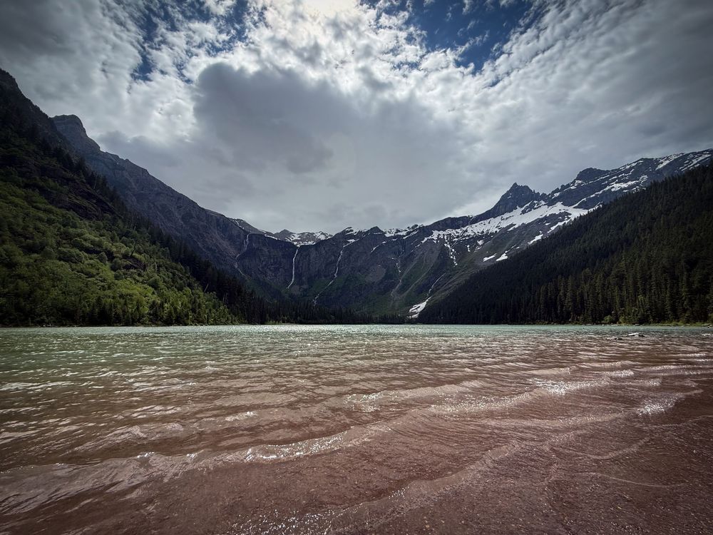 A serene mountain lake with sandy shores, snow-capped peaks, dense forests, and dramatic cloudy skies in a wilderness setting.
