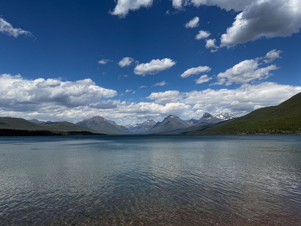 Serene mountain lake with crystal clear water reflecting blue skies and white clouds in Glacier National Park.
