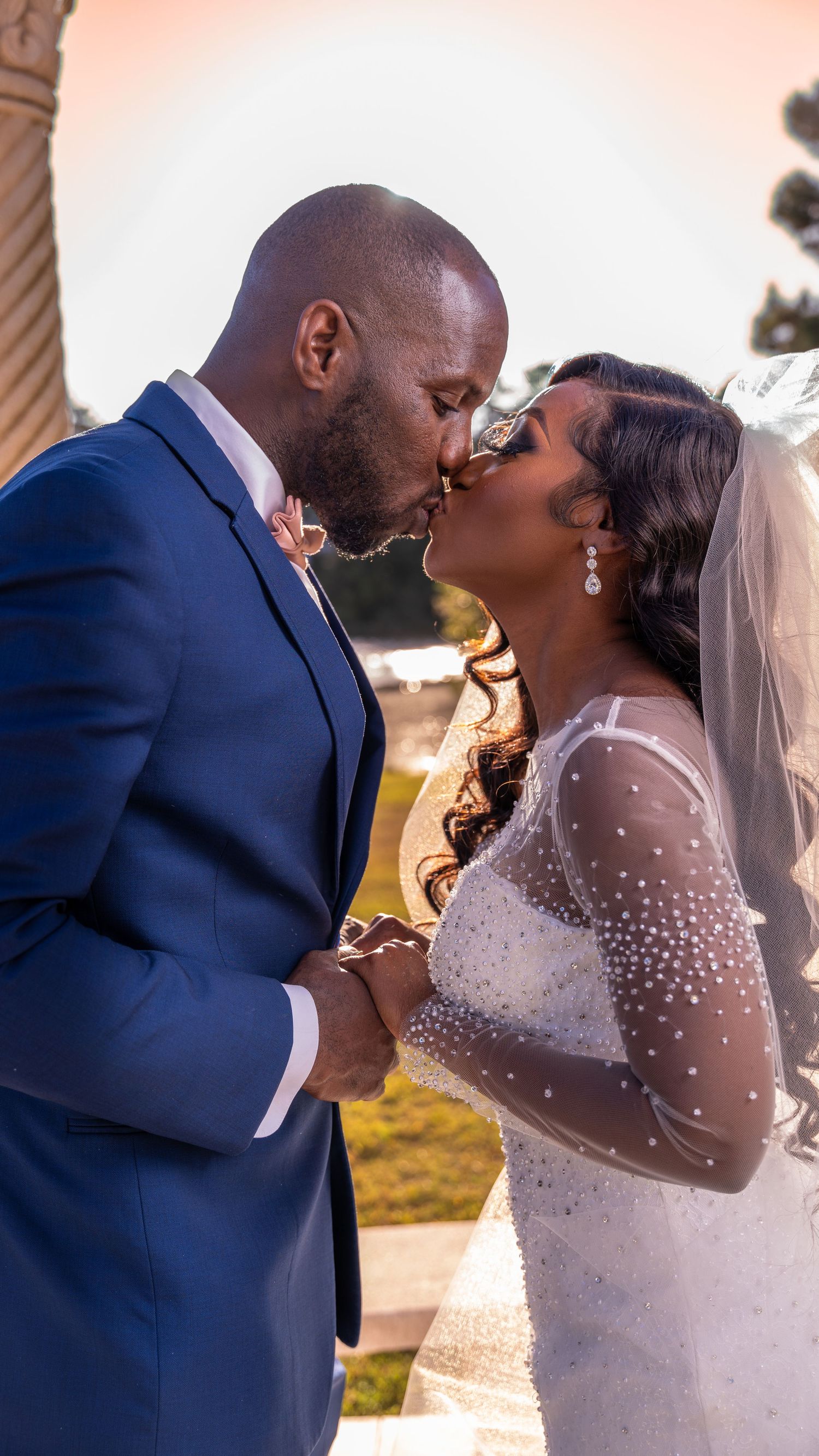A bride and groom sharing a romantic kiss at a beachside wedding, both wearing braided hairstyles, dressed in white lace and beige suit.