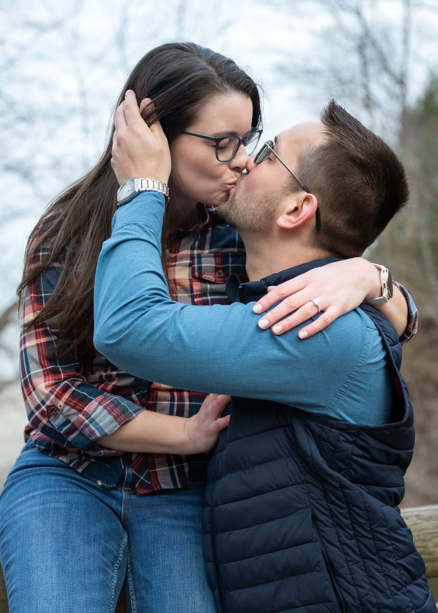 A couple sharing a romantic kiss outdoors, both wearing glasses and casual clothing, with the woman showing an engagement ring.