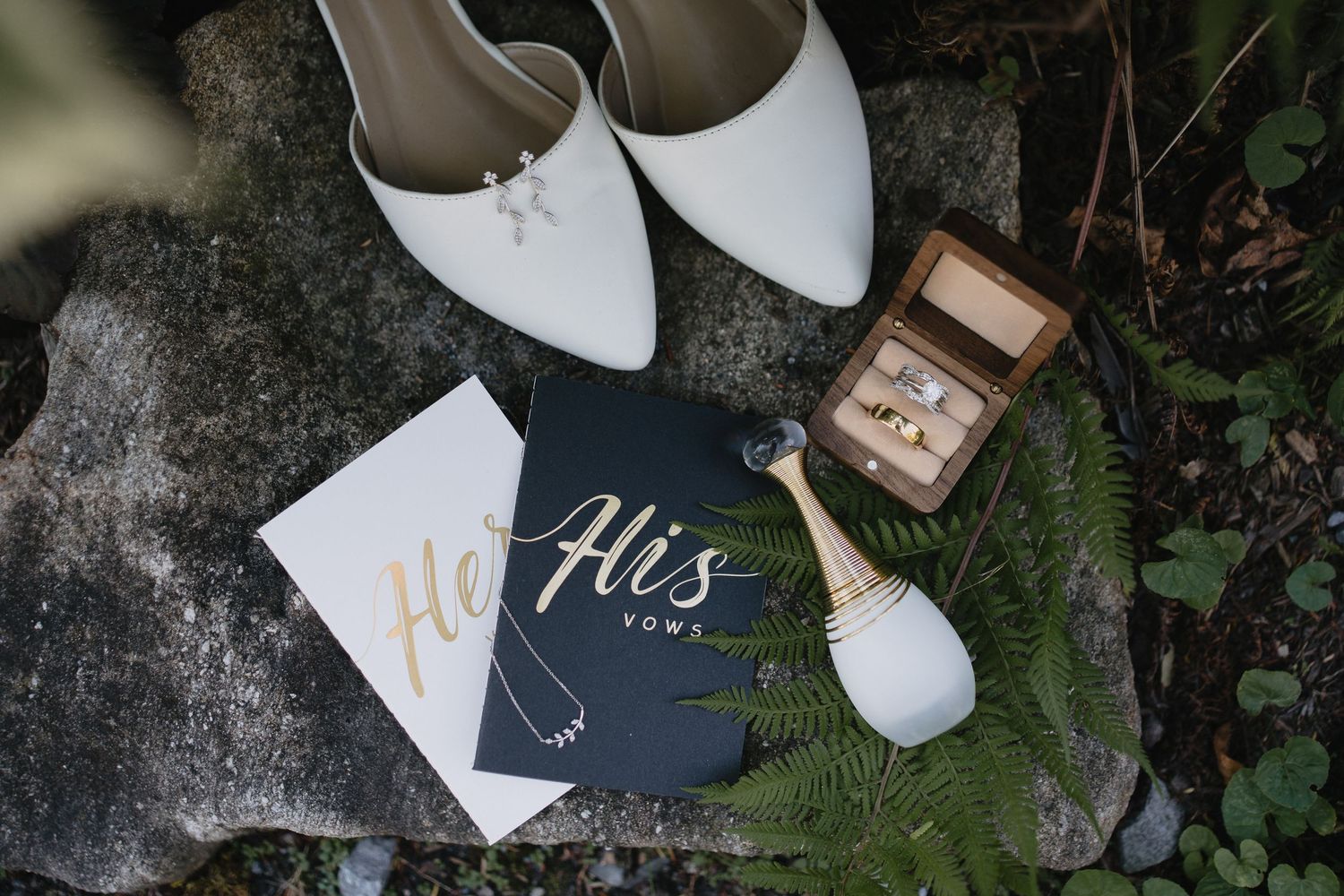 White bridal heels, His and Hers vow books, gold wedding rings in wooden box, and Dior perfume arranged on stone with ferns.