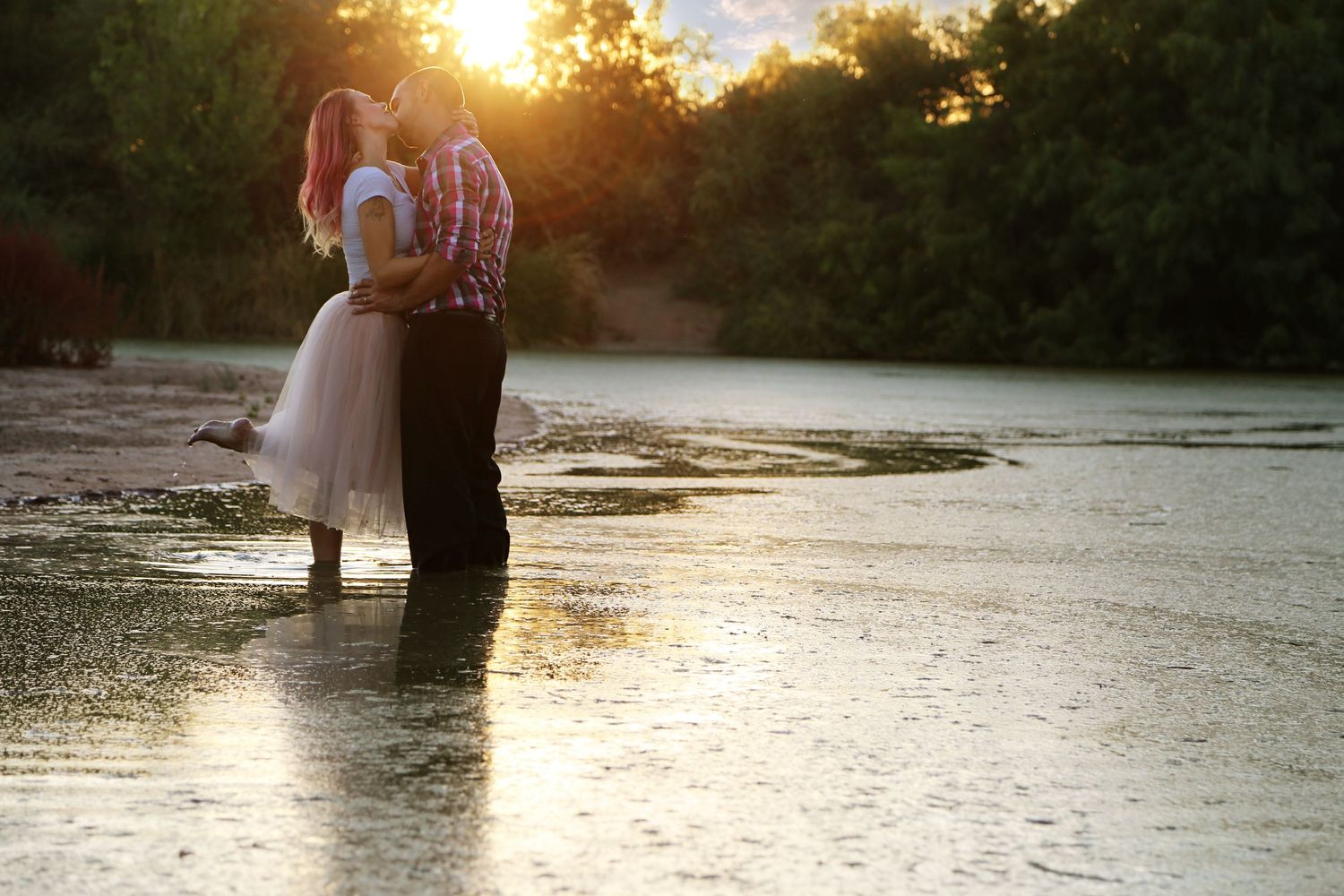 Couple in an embrace standing in shallow water at sunset, with golden sunlight glowing through the trees behind them.