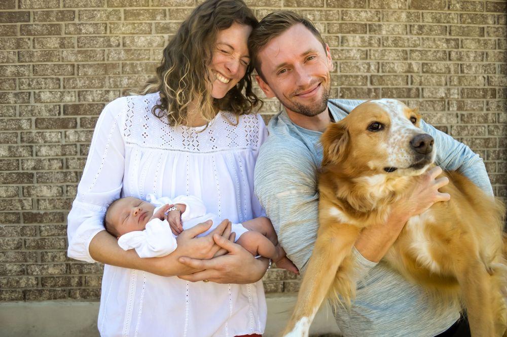 A young family poses for a picture, featuring mother, father, baby, and puppy