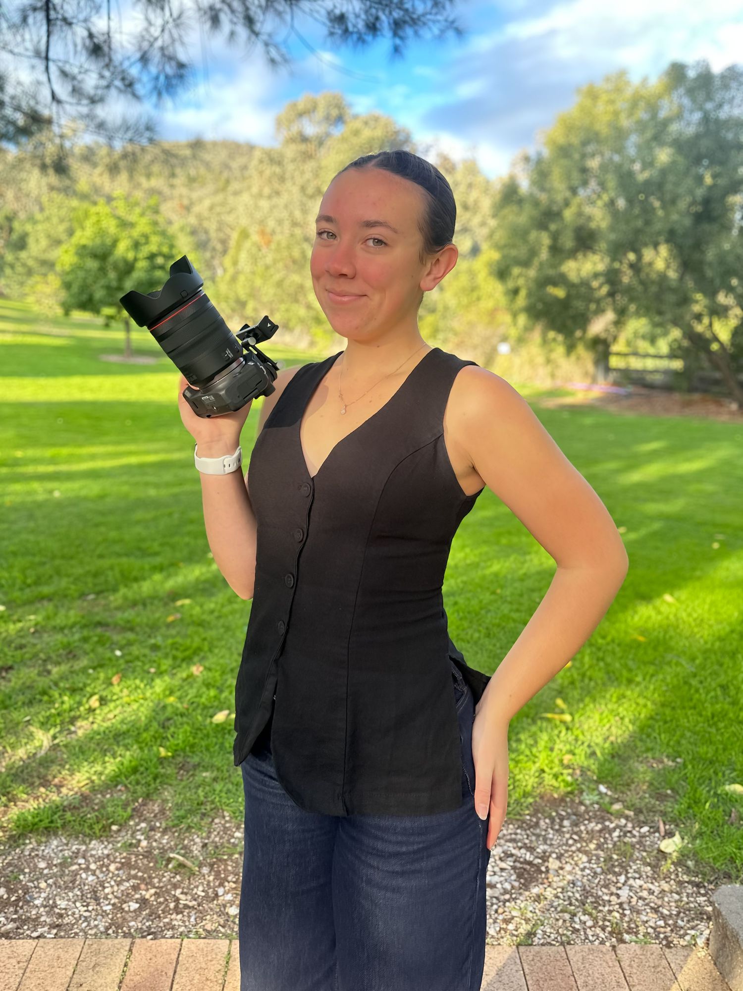 Woman in black vest holding a professional camera outdoors in a lush green park on a sunny day.