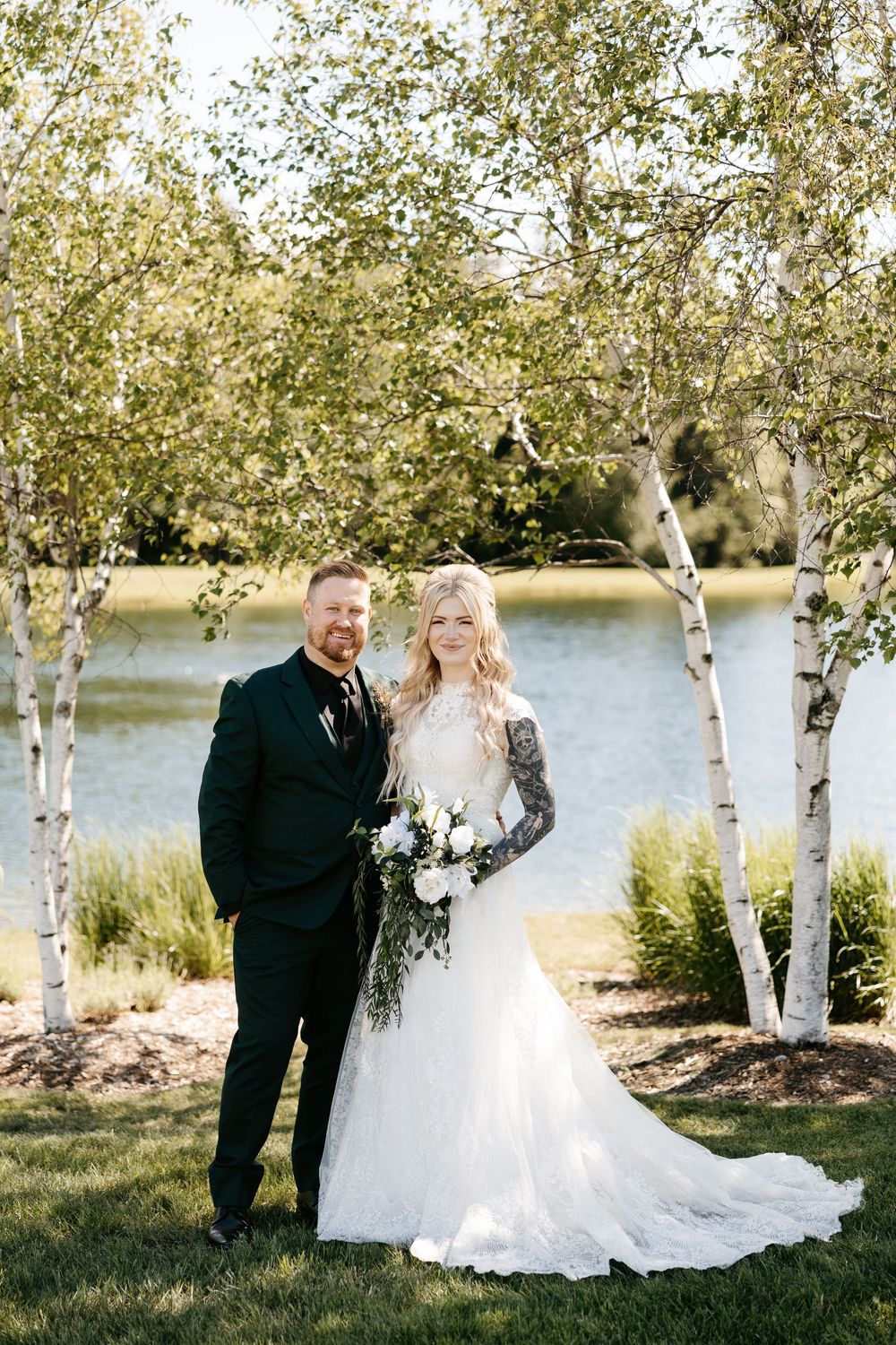 Bride and groom posing among birch trees near a lake, bride in lace gown holding white bouquet, groom in black suit.
