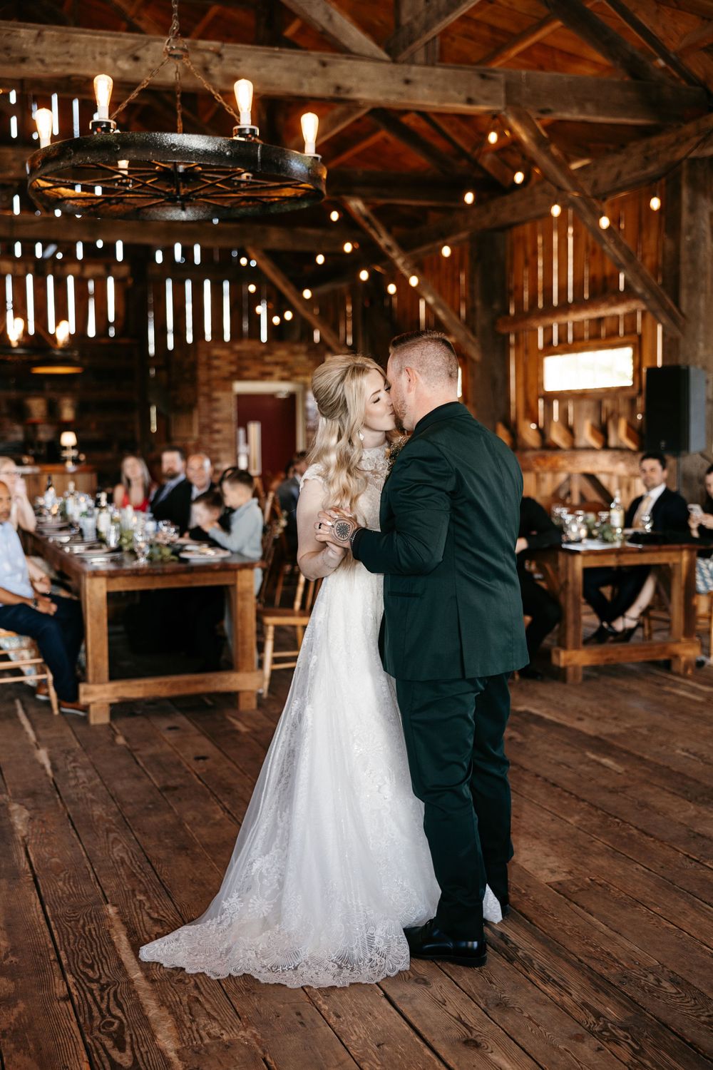 Bride and groom share first dance in a rustic barn wedding venue with string lights and wood beams.