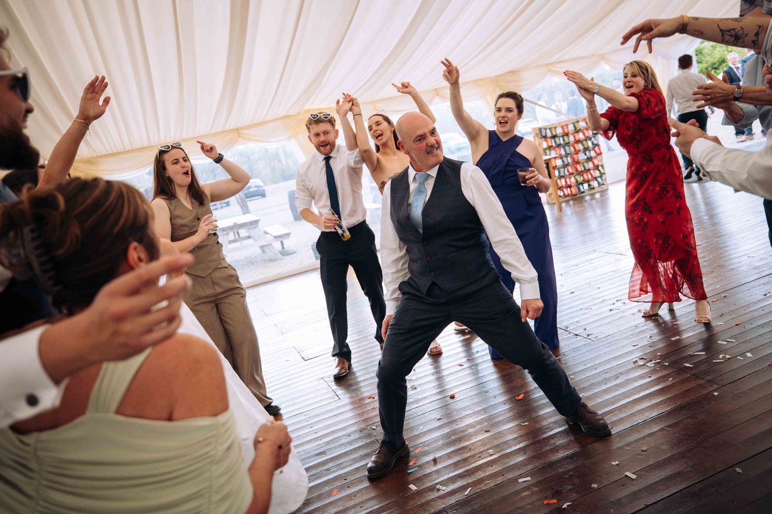 Outdoor event venue with white marquee tents, picnic tables, and guests gathered on a gravel patio under a cloudy sky.