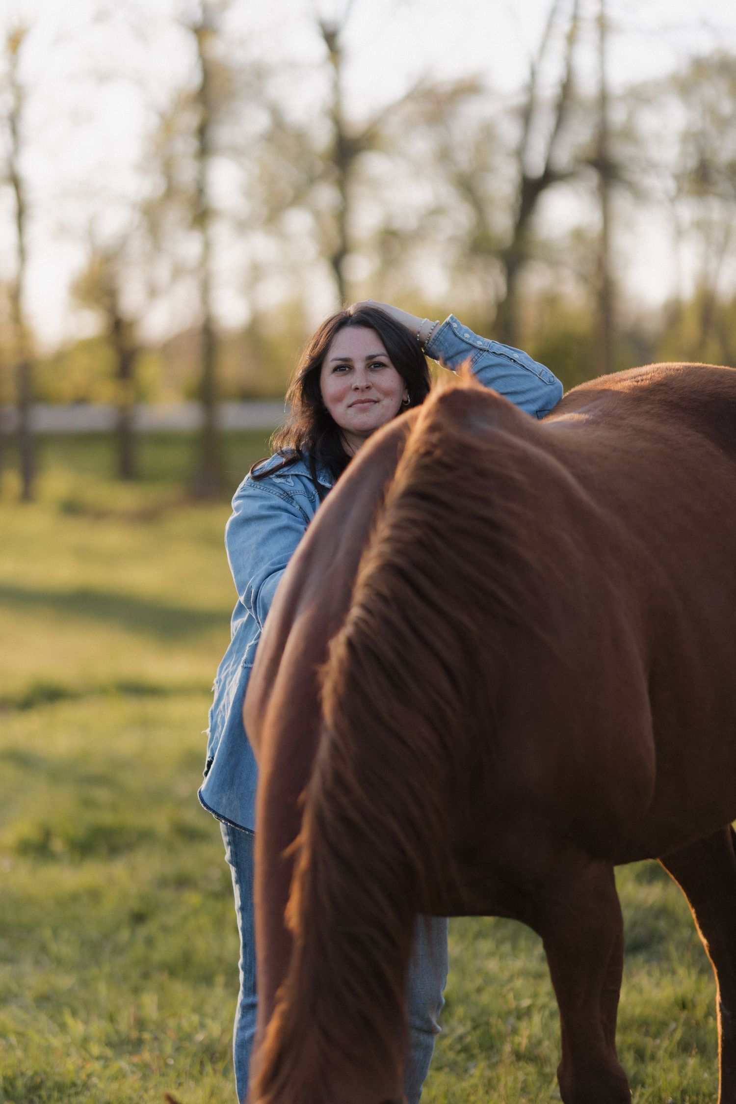 Smiling woman in denim jacket with stars petting a brown horse in a sunny green field.