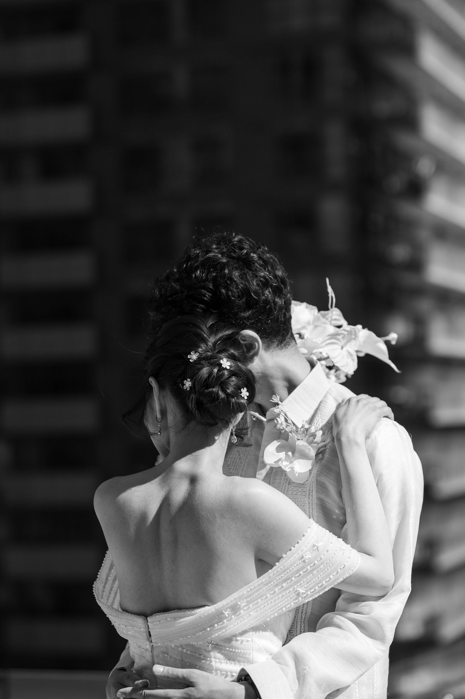 Black and white photo of a couple embracing, bride in strapless gown with floral hair accessories kissing her partner.