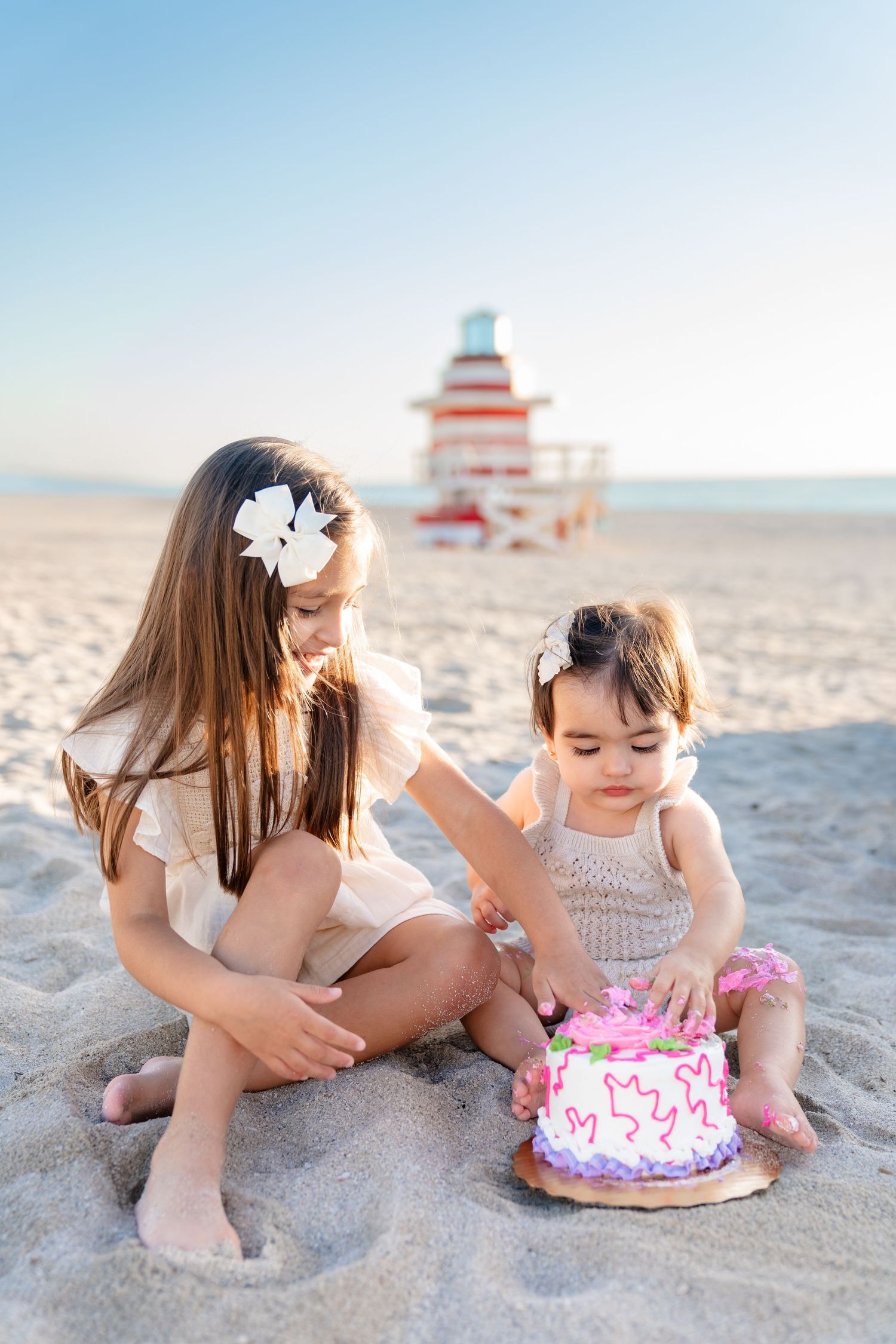 Baby in teal cap and pink shorts eating smash cake on a surfboard at the beach.