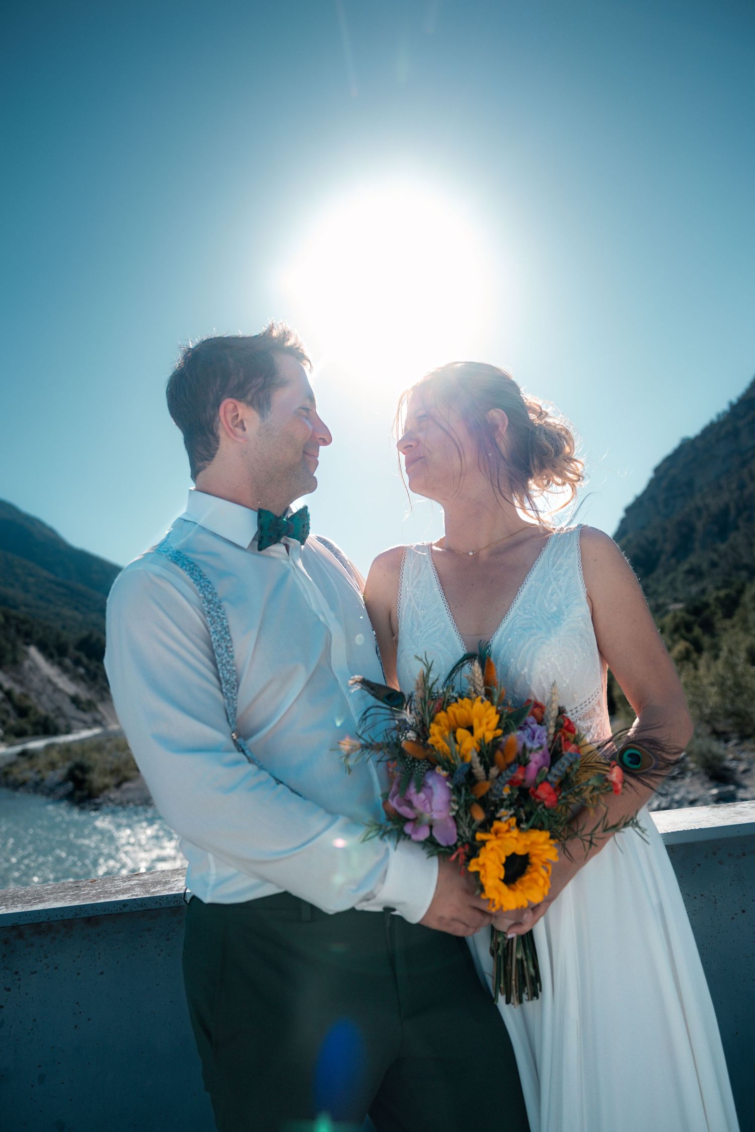 Bride and groom smiling at each other outdoors, holding a sunflower bouquet with mountains and bright sun behind them.