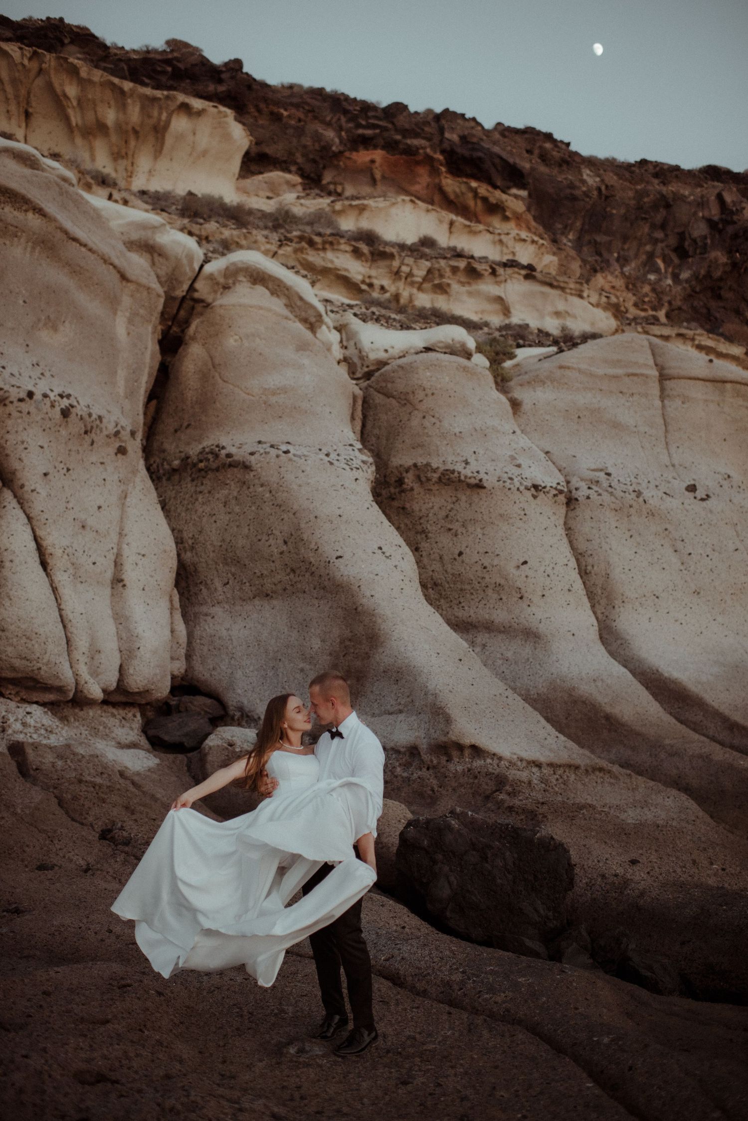 Couple in wedding attire sharing a romantic kiss against dramatic white rock formations under a moonlit desert sky.