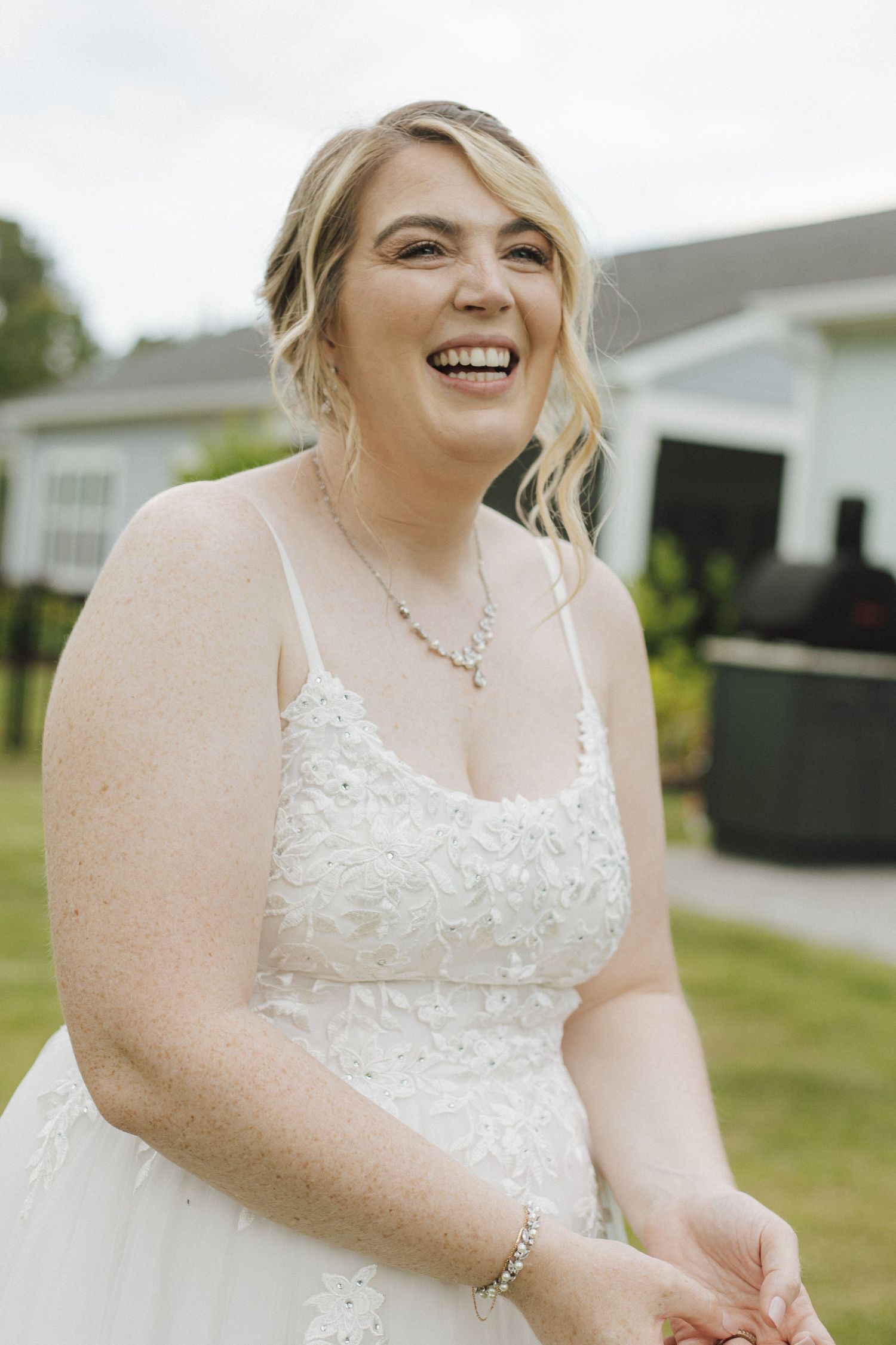 Smiling bride in white lace wedding gown with spaghetti straps and delicate necklace laughing outdoors on her wedding day.