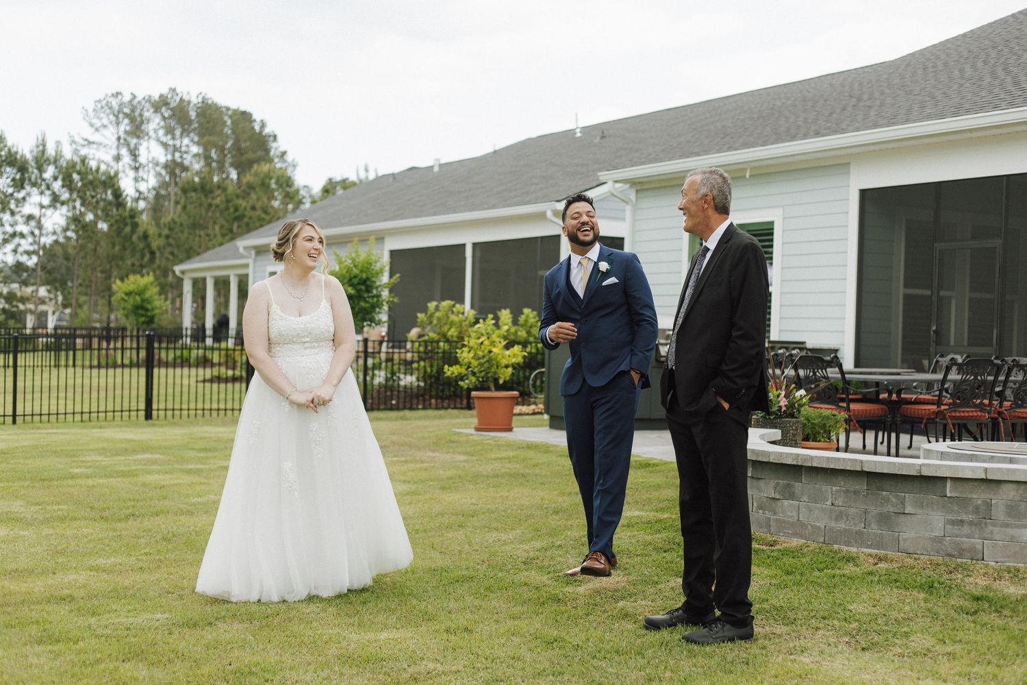 Bride in white gown and groom in navy suit stand with officiant on green lawn during outdoor wedding ceremony.