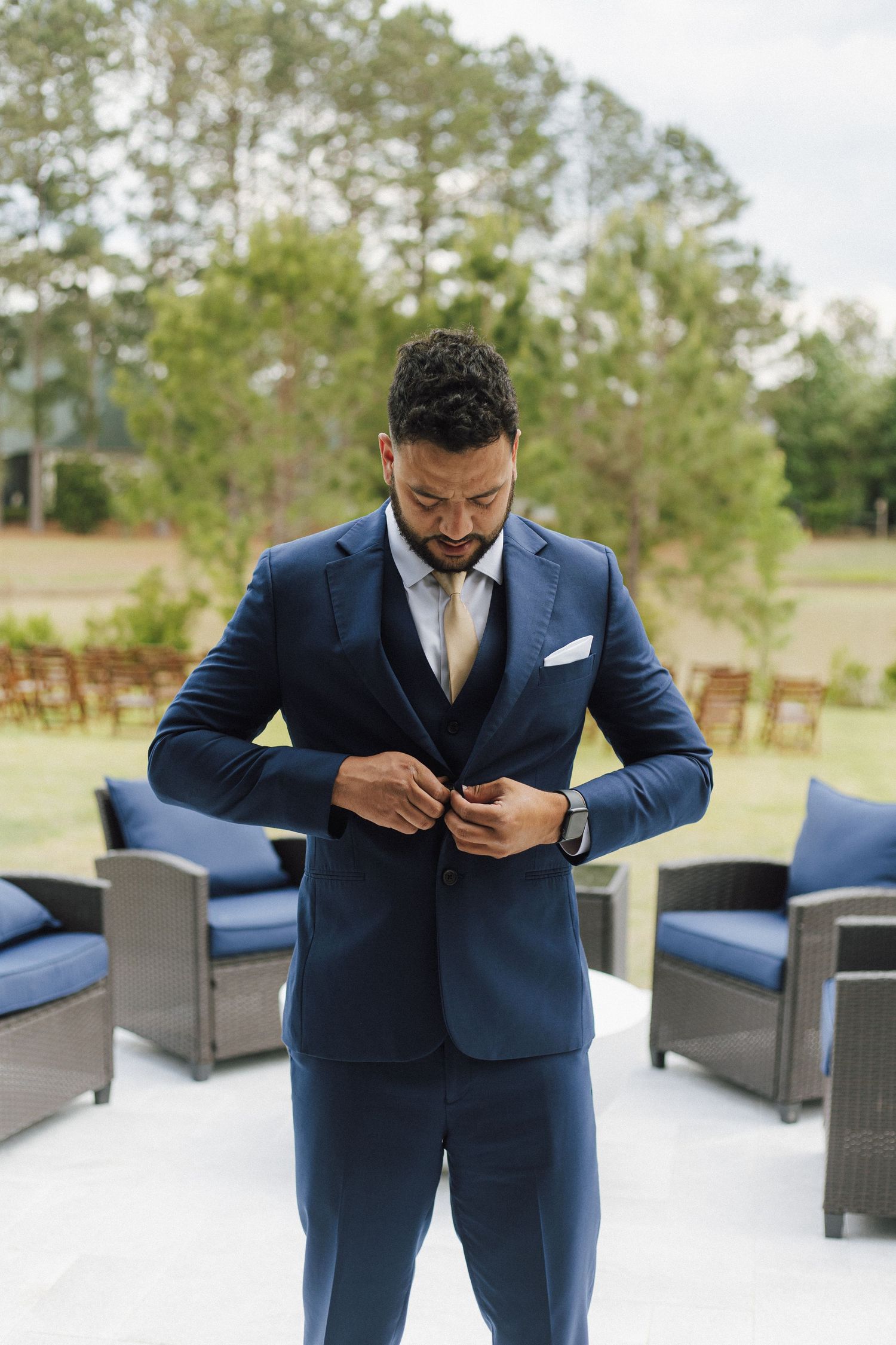 A groom in a navy blue three-piece suit adjusts his jacket button outdoors at a wedding venue.