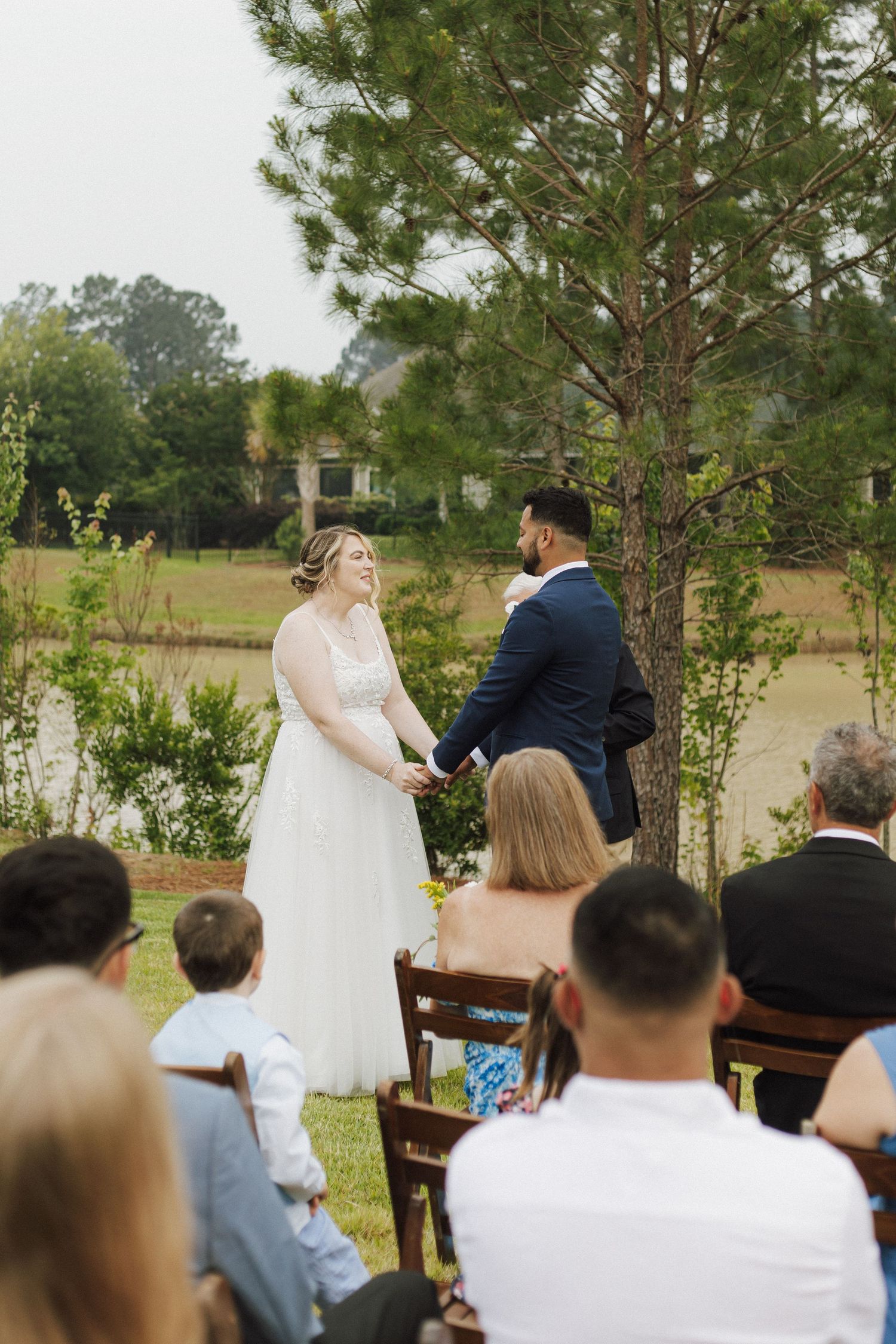 Bride and groom hold hands during outdoor wedding ceremony near a pond, with seated guests watching in a lush green setting.
