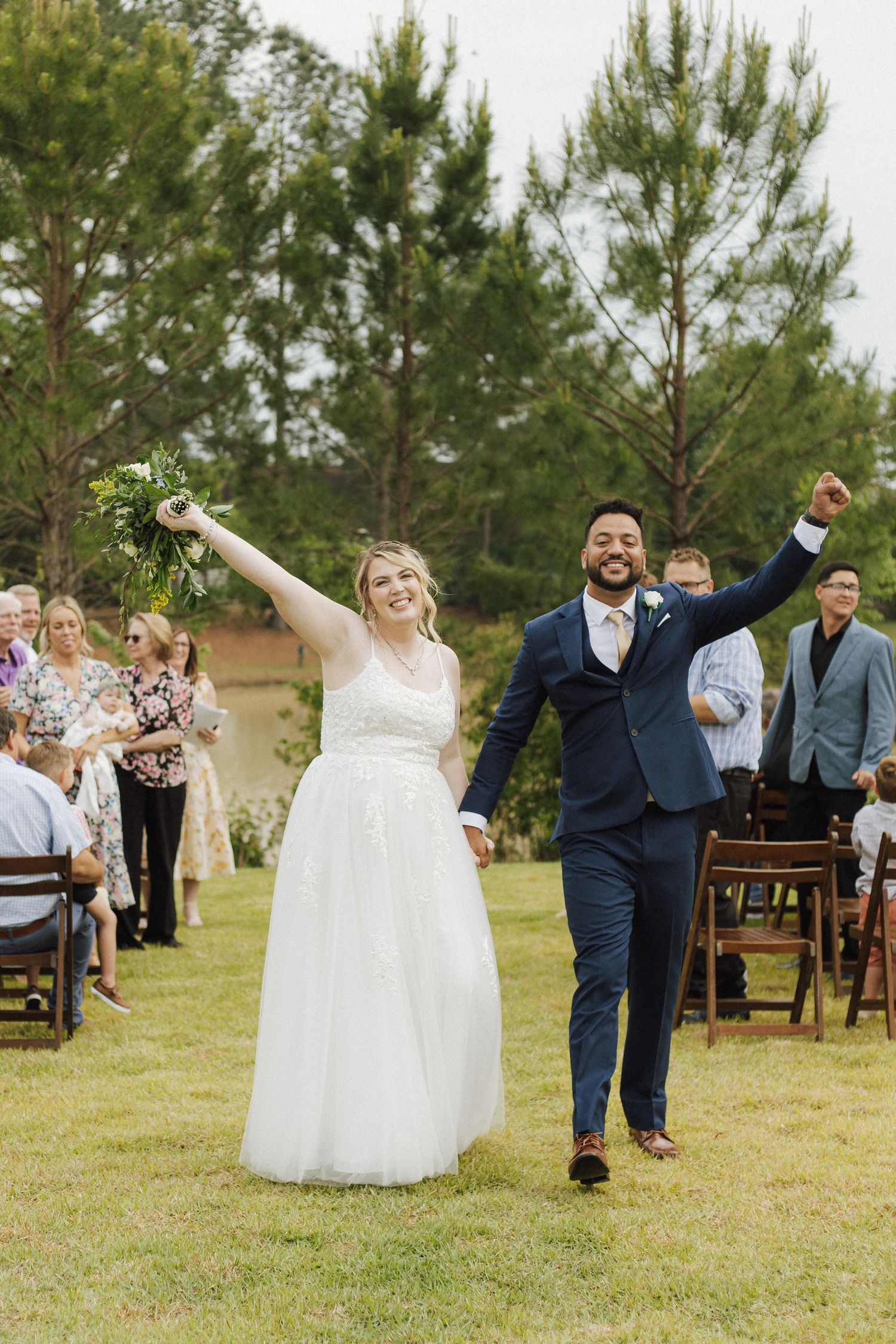 Joyful newlyweds celebrate after outdoor wedding ceremony, bride in white gown holding bouquet, groom in navy suit, raising hands.