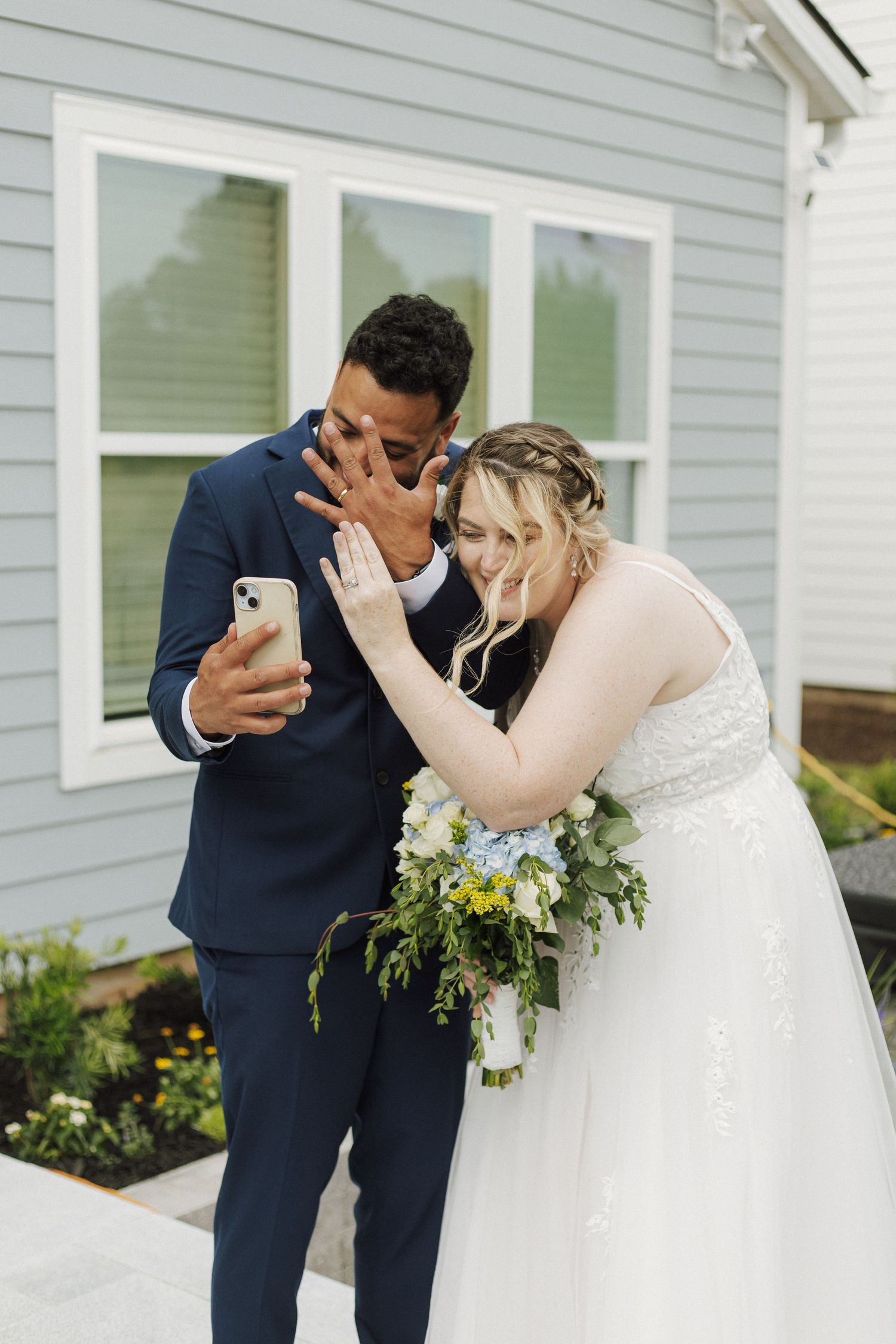 Bride and groom laughing while taking a selfie outdoors, bride holding a bouquet of white and yellow flowers.