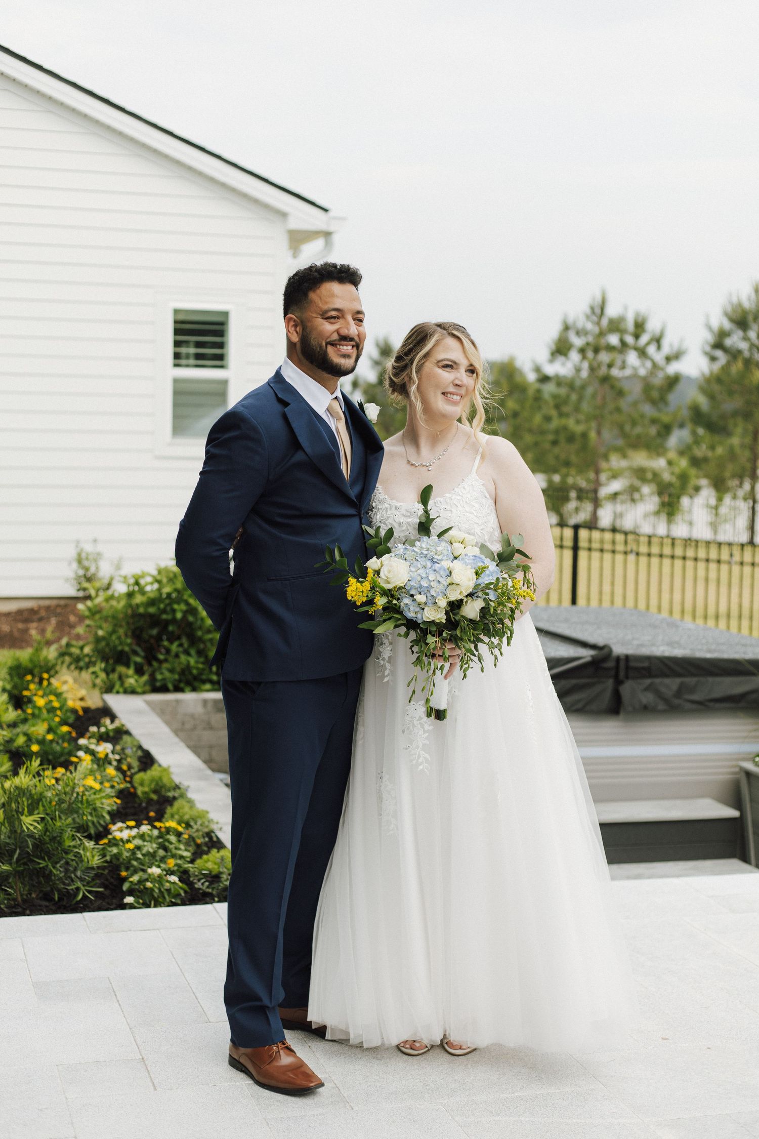 Bride and groom posing outdoors on patio, groom in navy suit, bride in white gown holding blue and yellow bouquet.