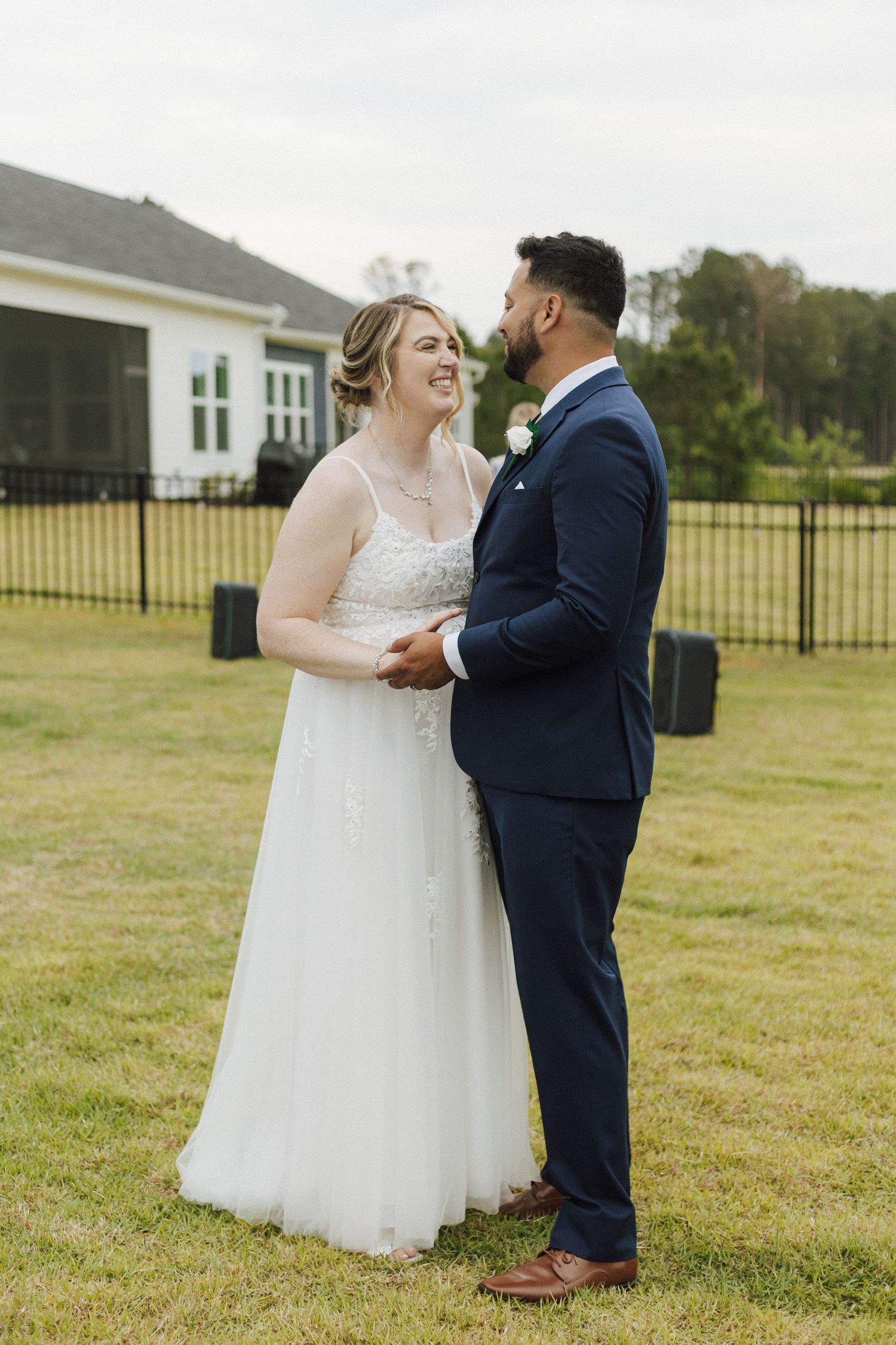 A bride in a white gown and groom in a navy suit hold hands and smile at each other outdoors on a green lawn.