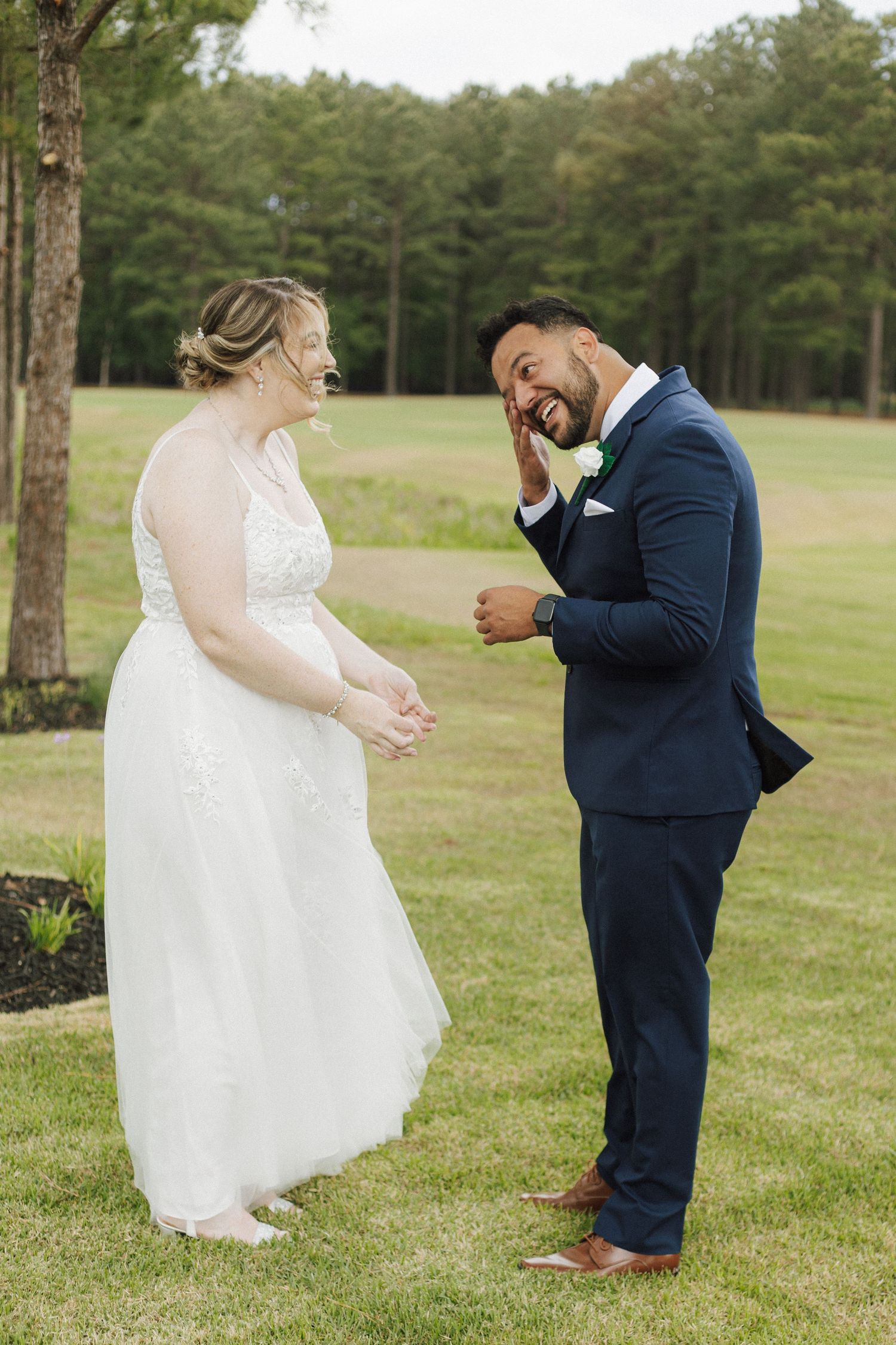 A bride in a white gown and groom in a navy suit share a joyful first look moment on a green lawn.