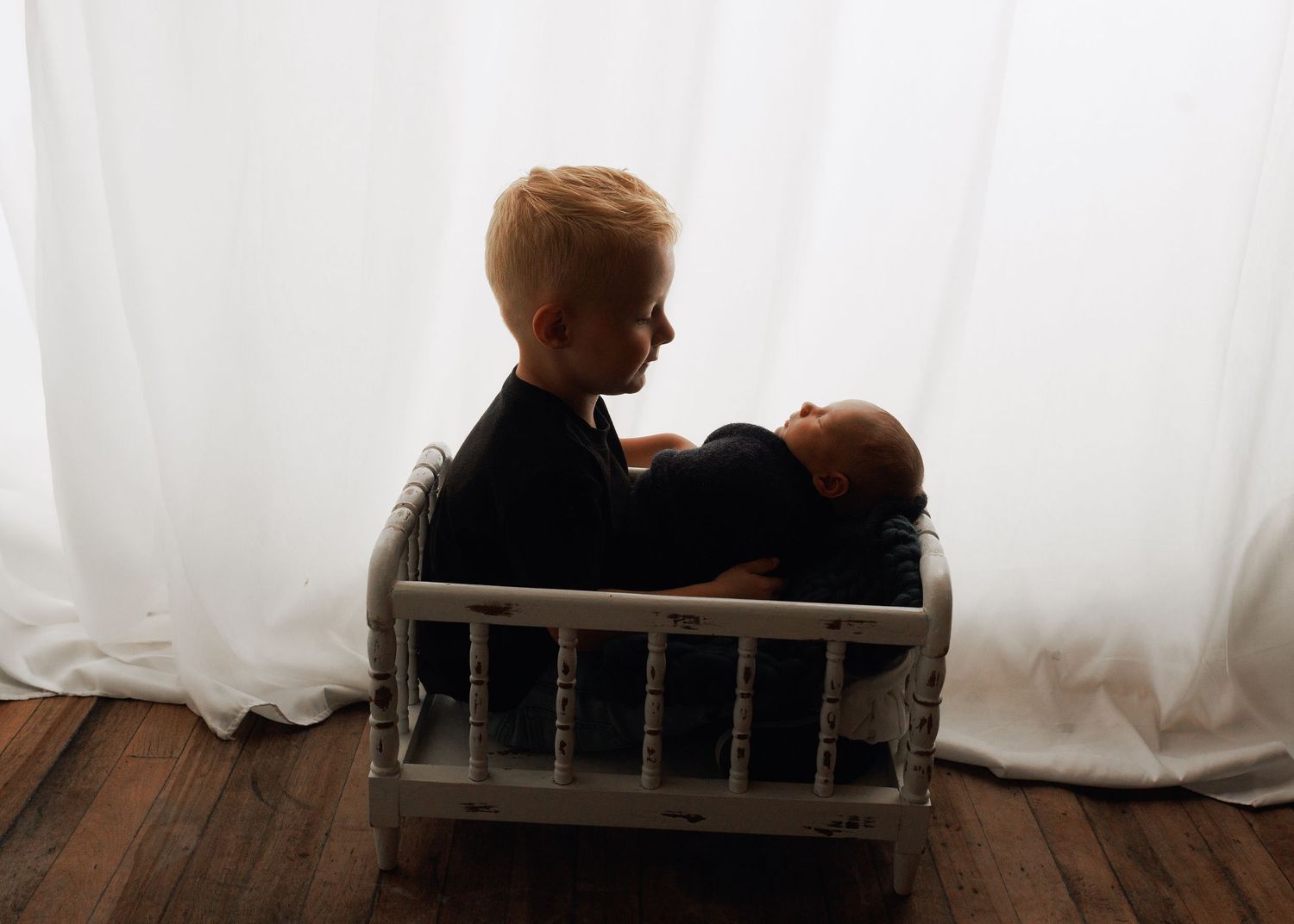 Older boy sitting with newborn baby in a small white crib, silhouetted against white curtains.