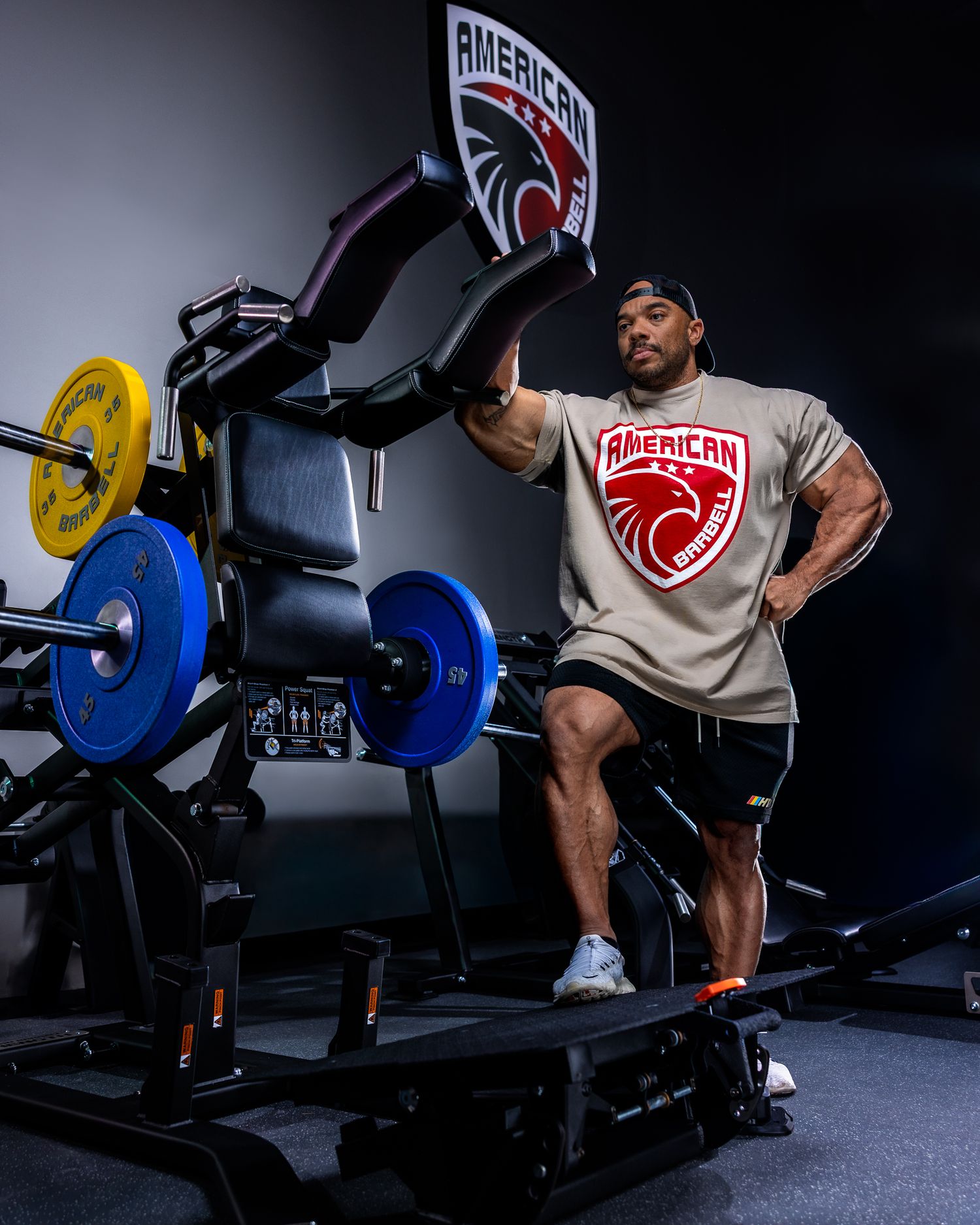 Muscular man in American Strength t-shirt posing beside a loaded leg press machine in a gym.