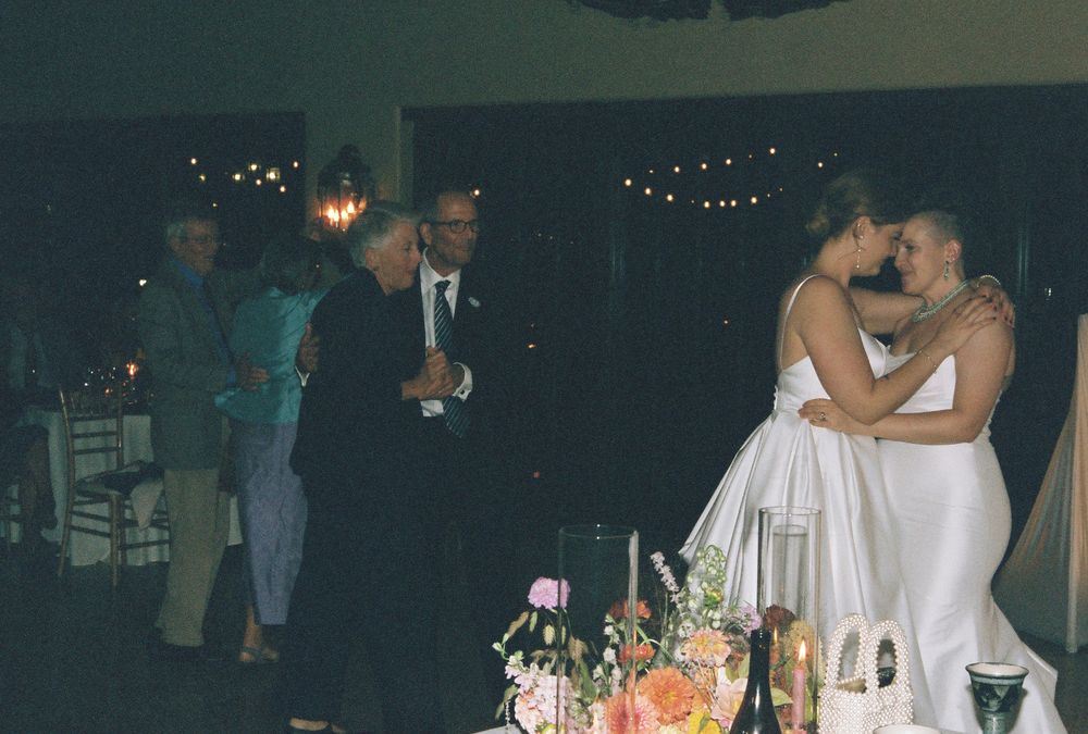 Two brides share a first dance kiss at their wedding reception while guests look on in a dimly lit venue.
