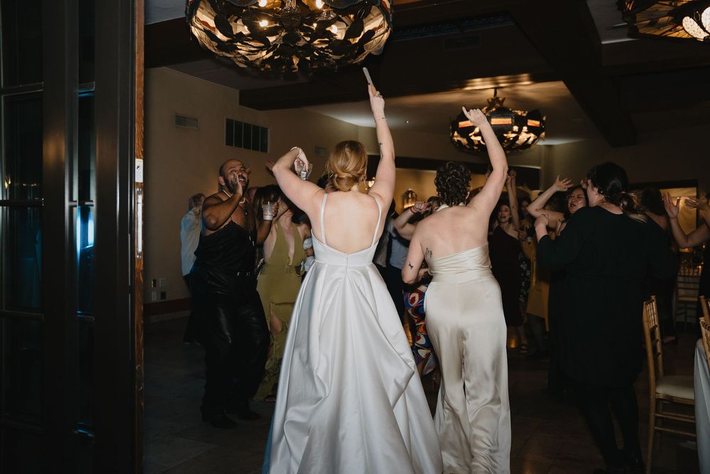 Two brides in white gowns celebrate with raised arms on a wedding reception dance floor.