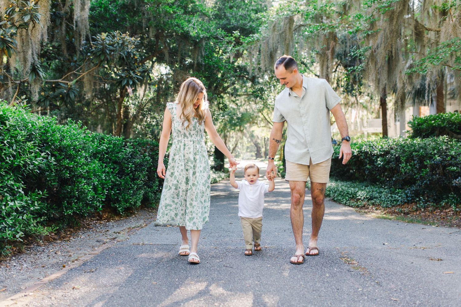 A young family in Beaufort walks hand-in-hand down a mossy oak tree-lined path, parents swinging their toddler between them.
