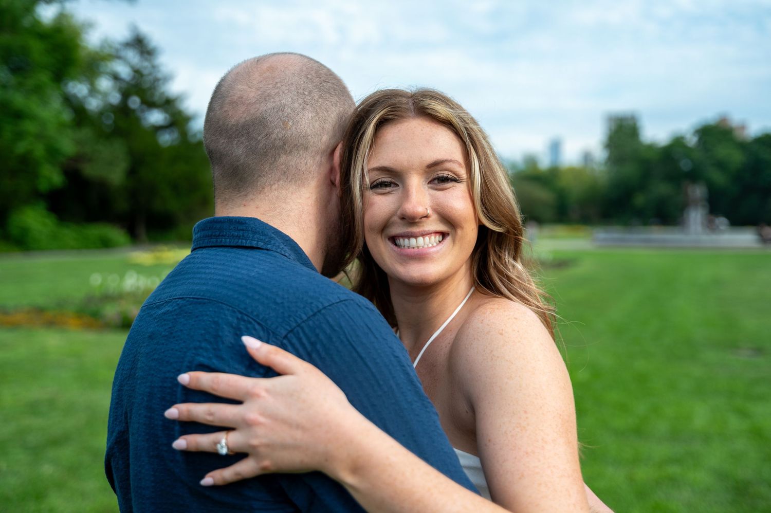 A smiling bride embraces her partner outdoors in a lush green park, showcasing her engagement ring.