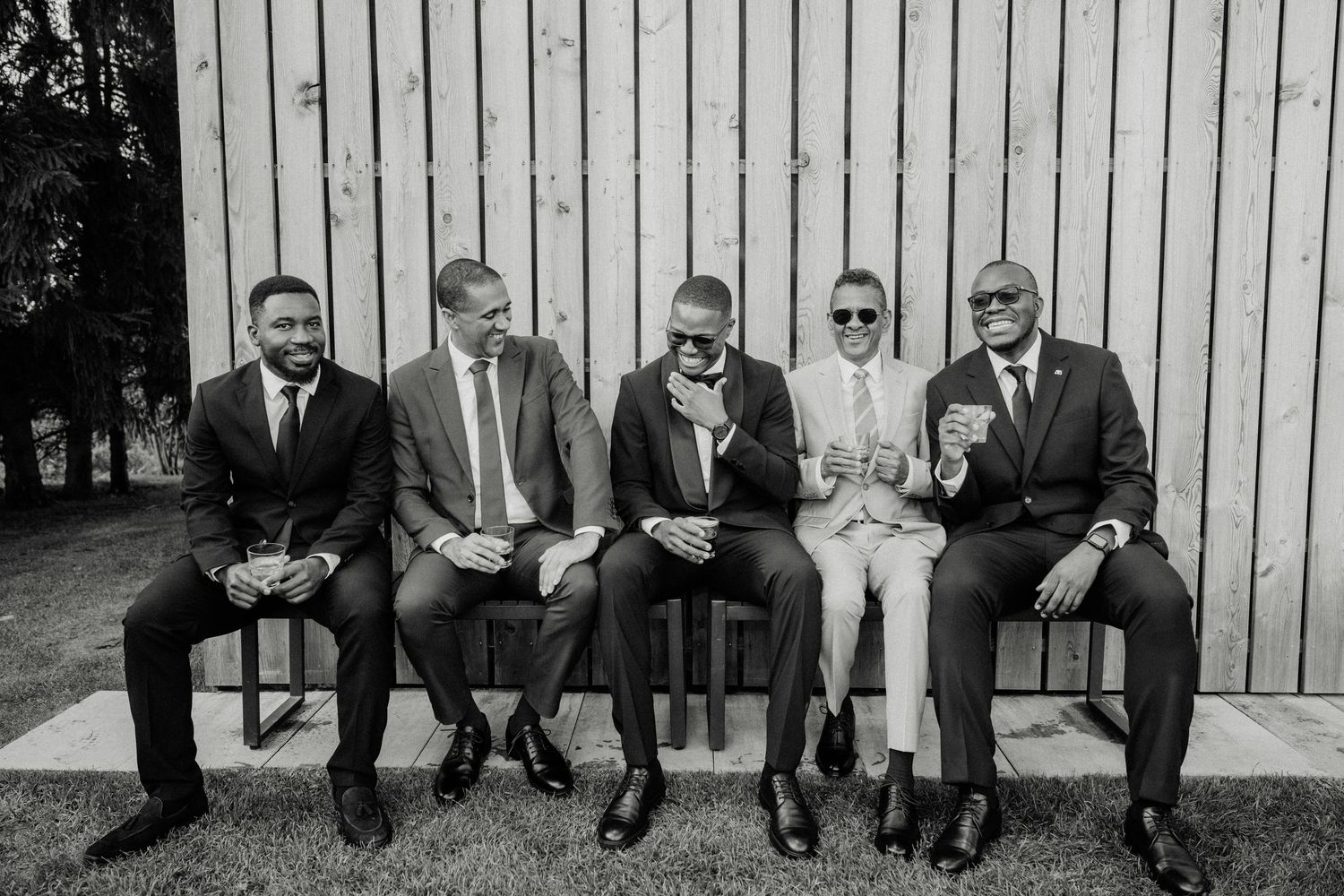 Five men in suits sitting on a bench laughing together in a black and white outdoor photo.