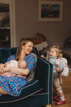 Mother relaxing on blue couch with baby while toddler plays nearby.
