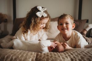 Children in white outfits sharing sweet moment while meeting their new sibling on bed.