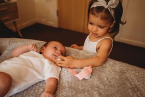 Young sister tenderly interacts with baby sibling lying on grey bedding.