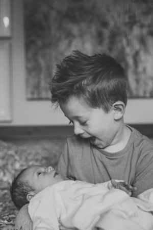 A black and white portrait of two siblings sharing a tender moment as the older one holds a newborn baby.