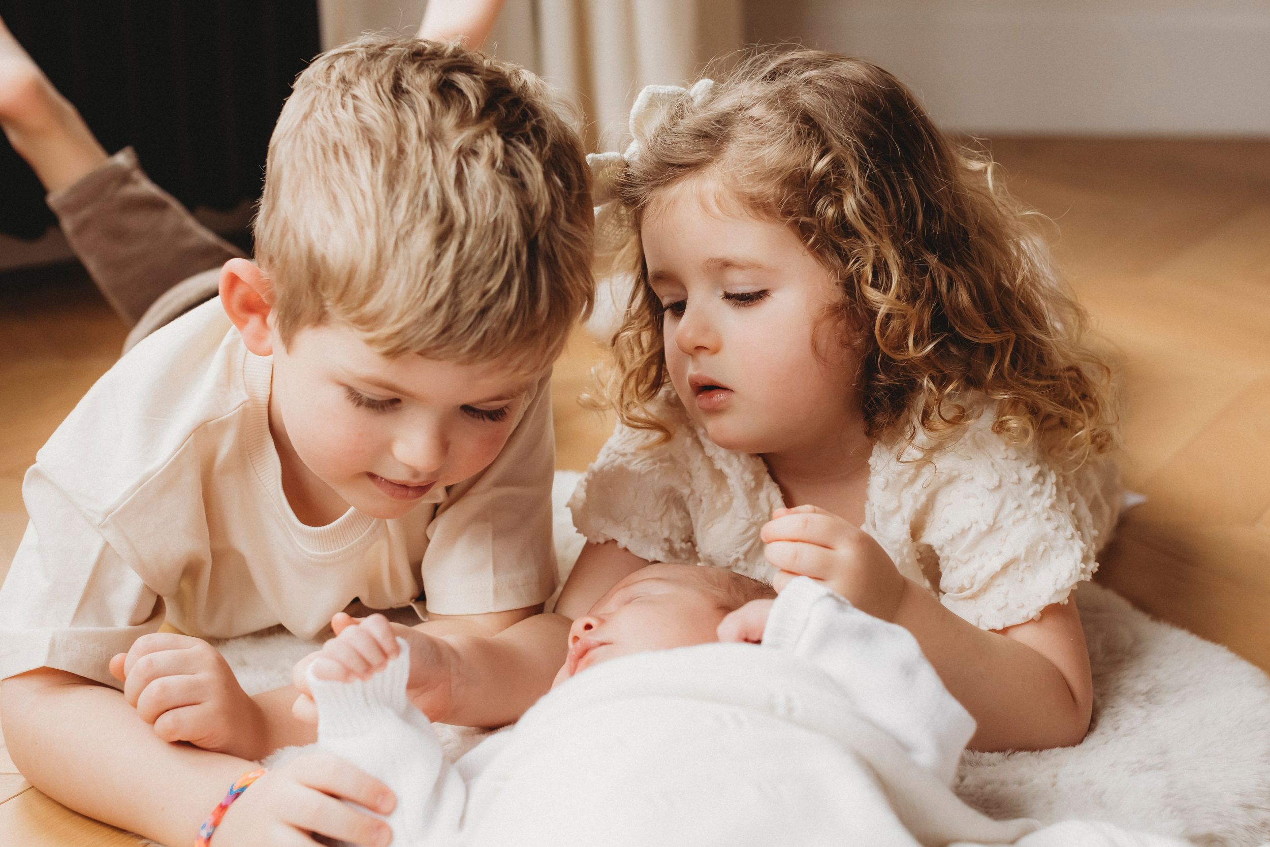 Young siblings leaning in close to look at newborn baby lying between them.