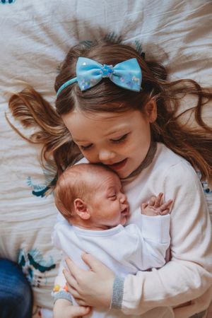 A sweet moment as a sibling with a blue bow gently cuddles their newborn baby on white bedding.