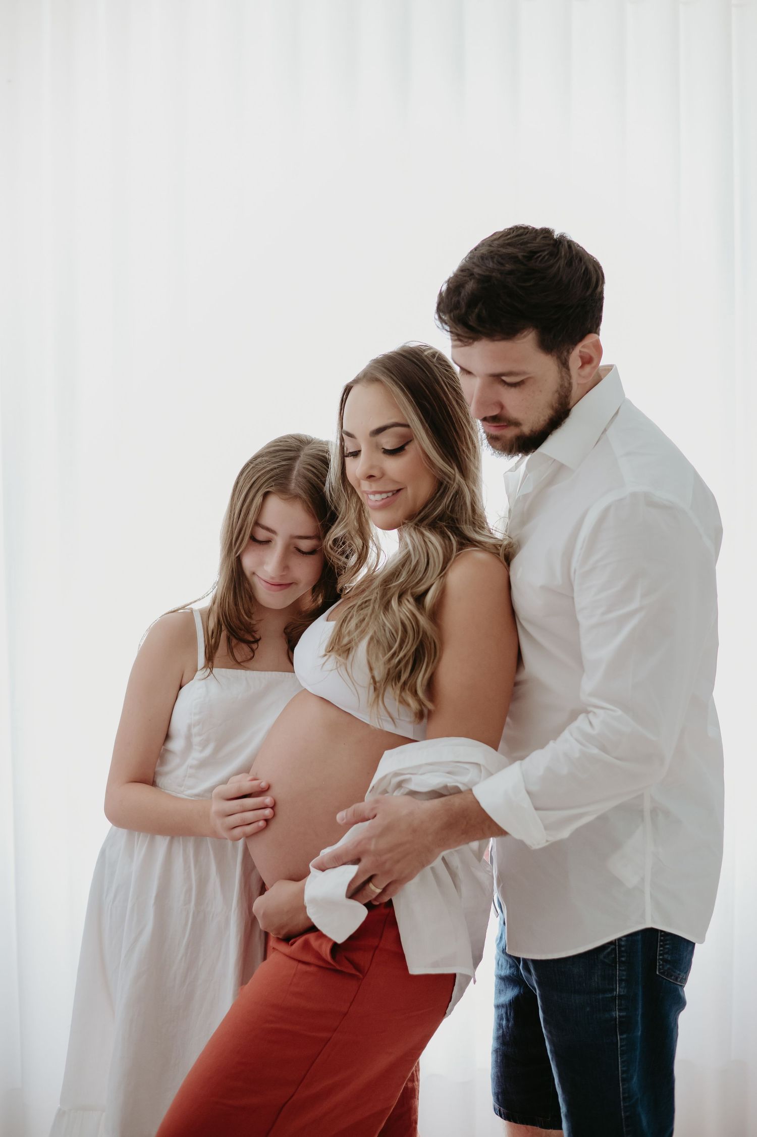 Pregnant woman in white bralette poses with partner and child holding baby shoes against her belly.