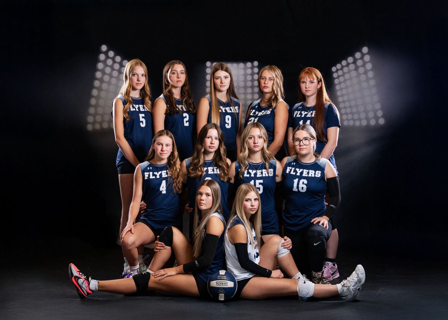 Girls volleyball team in navy Flyers uniforms posing for official team photo in front of stadium lights backdrop.