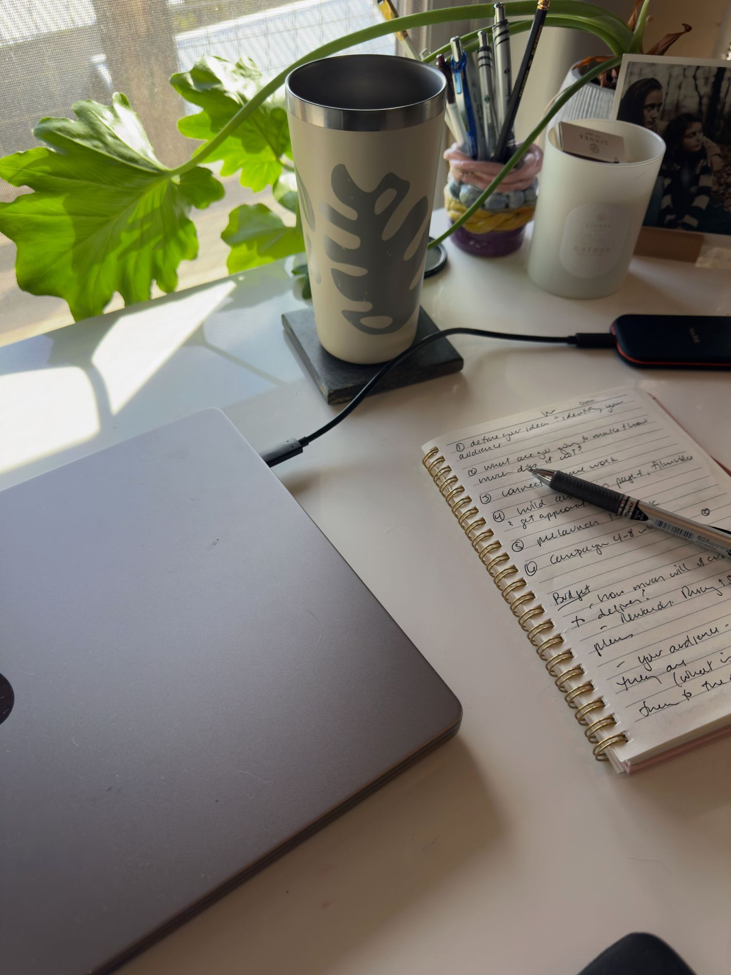 A cozy desk workspace with a MacBook, spiral notebook, pen, travel mug, plant, and desk organizer in natural sunlight in Asheville, NC
