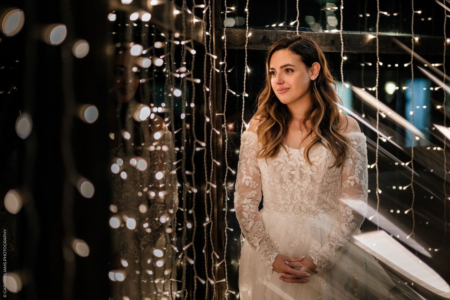Bride in lace wedding gown standing among sparkling fairy lights in a dark, romantic setting.