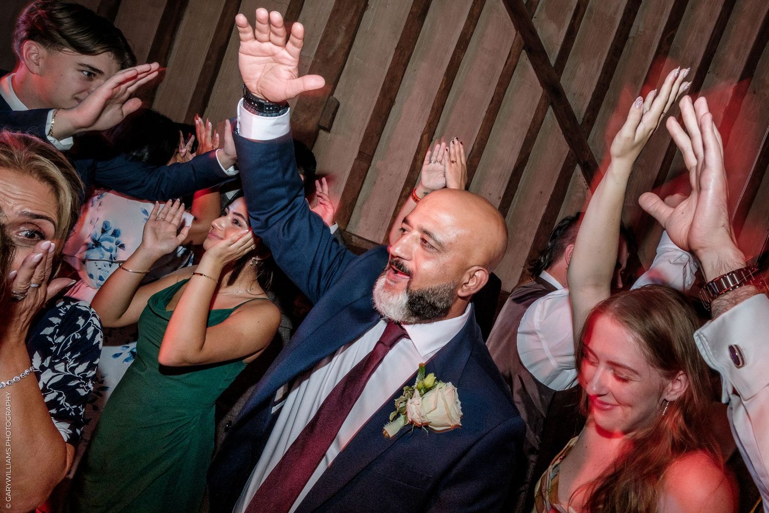 Bald groom in suit raises hand joyfully on wedding dance floor surrounded by celebrating guests.