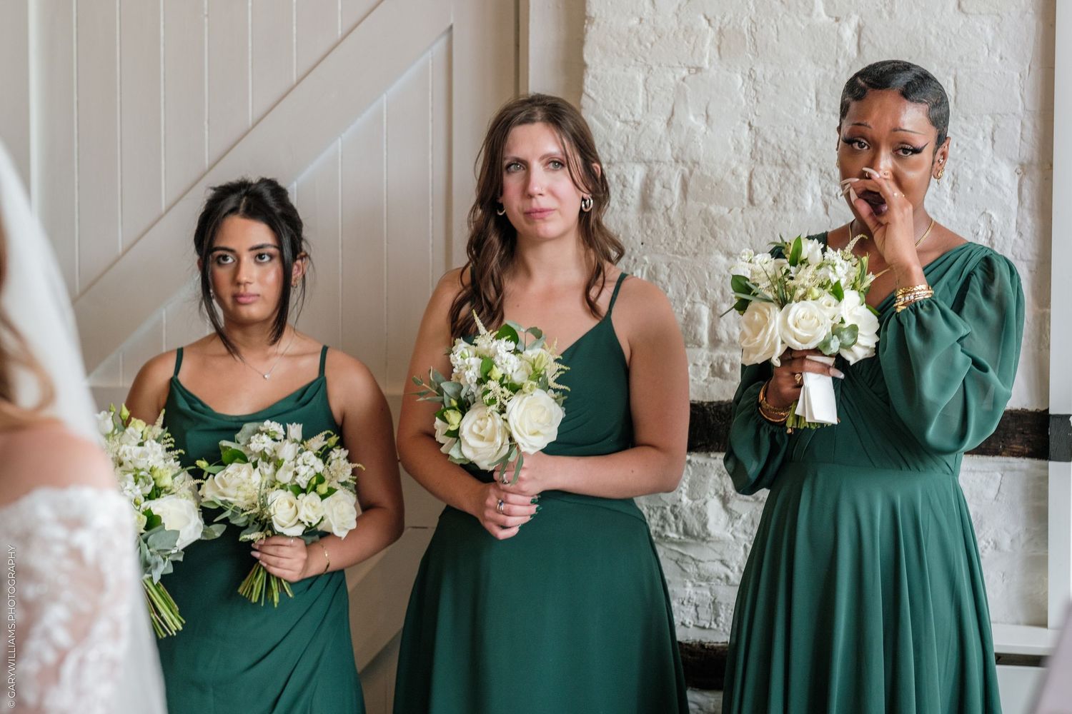 Three bridesmaids in emerald green dresses holding white bouquets react emotionally while watching a bride during a wedding ceremony.