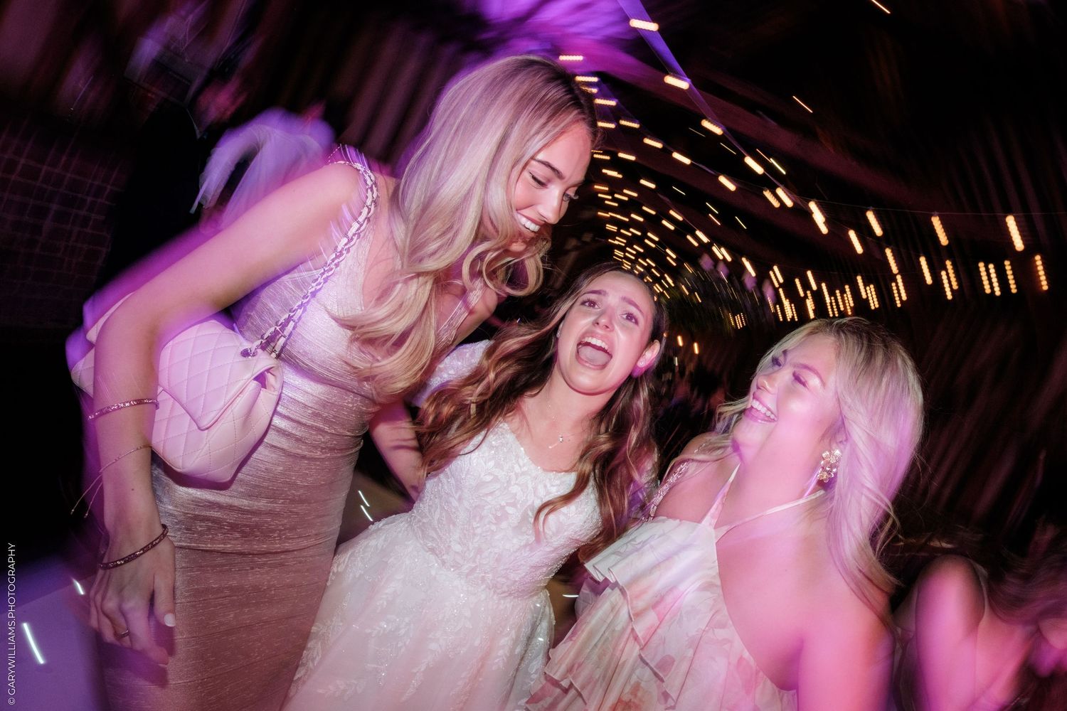 Three women laughing and dancing joyfully at a wedding reception under purple lights.