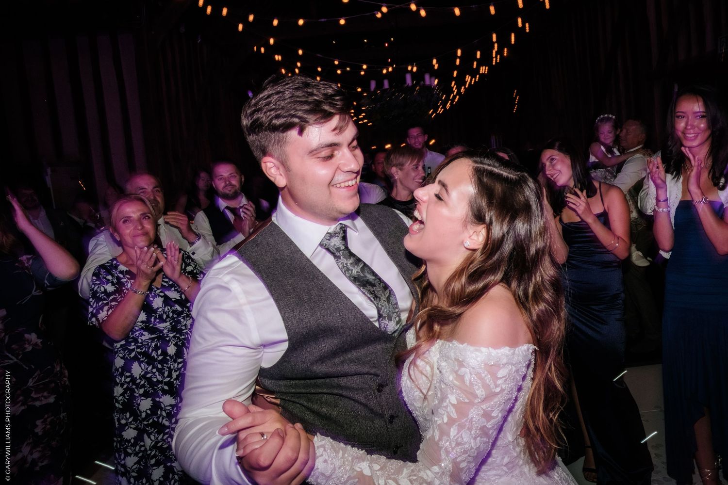 Bride and groom share a joyful first dance surrounded by cheering wedding guests in a warmly lit reception venue.